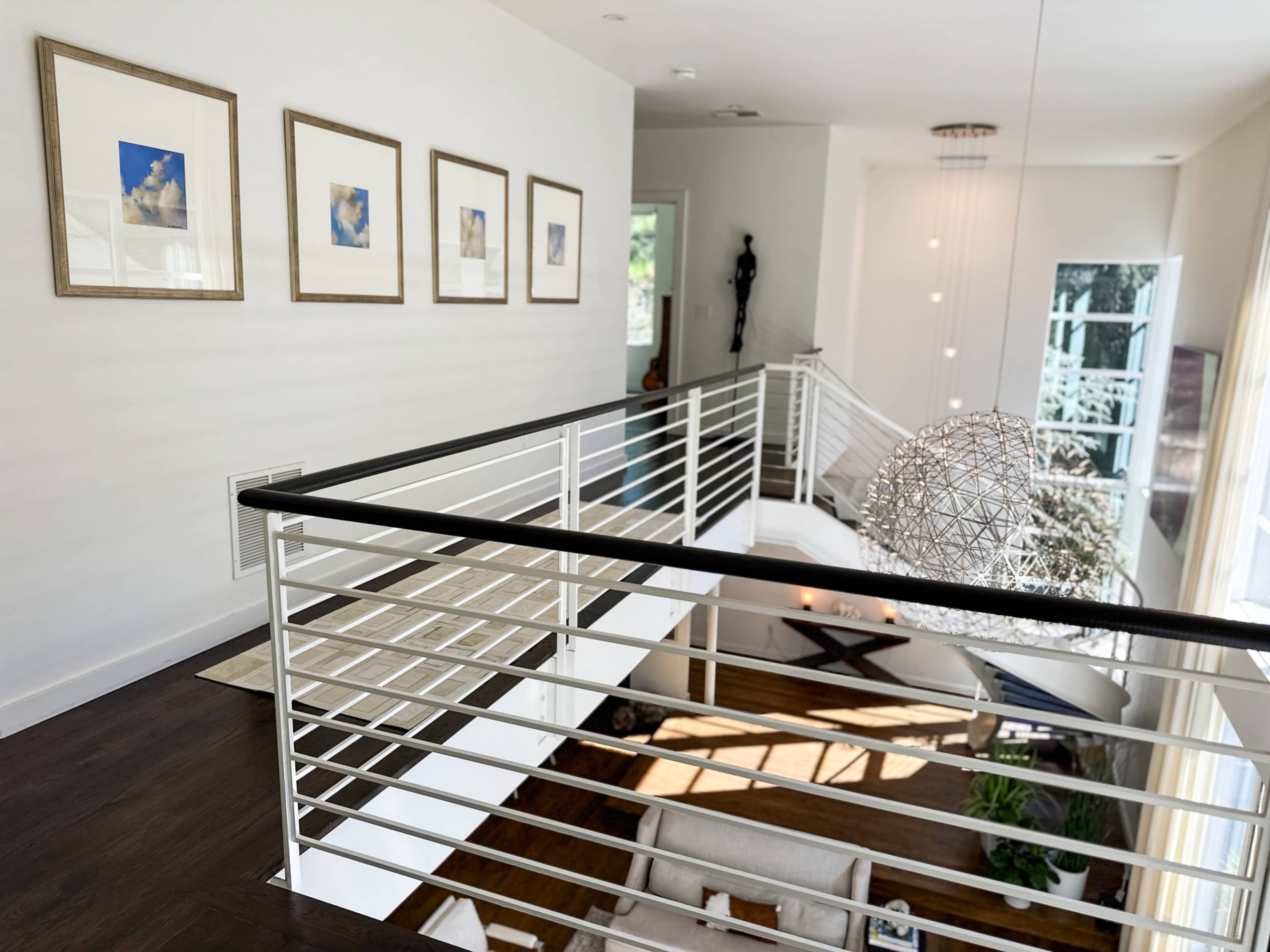 The image shows a modern interior with a balcony overlooking a living area, featuring framed pictures on the wall and a geometric light fixture above.