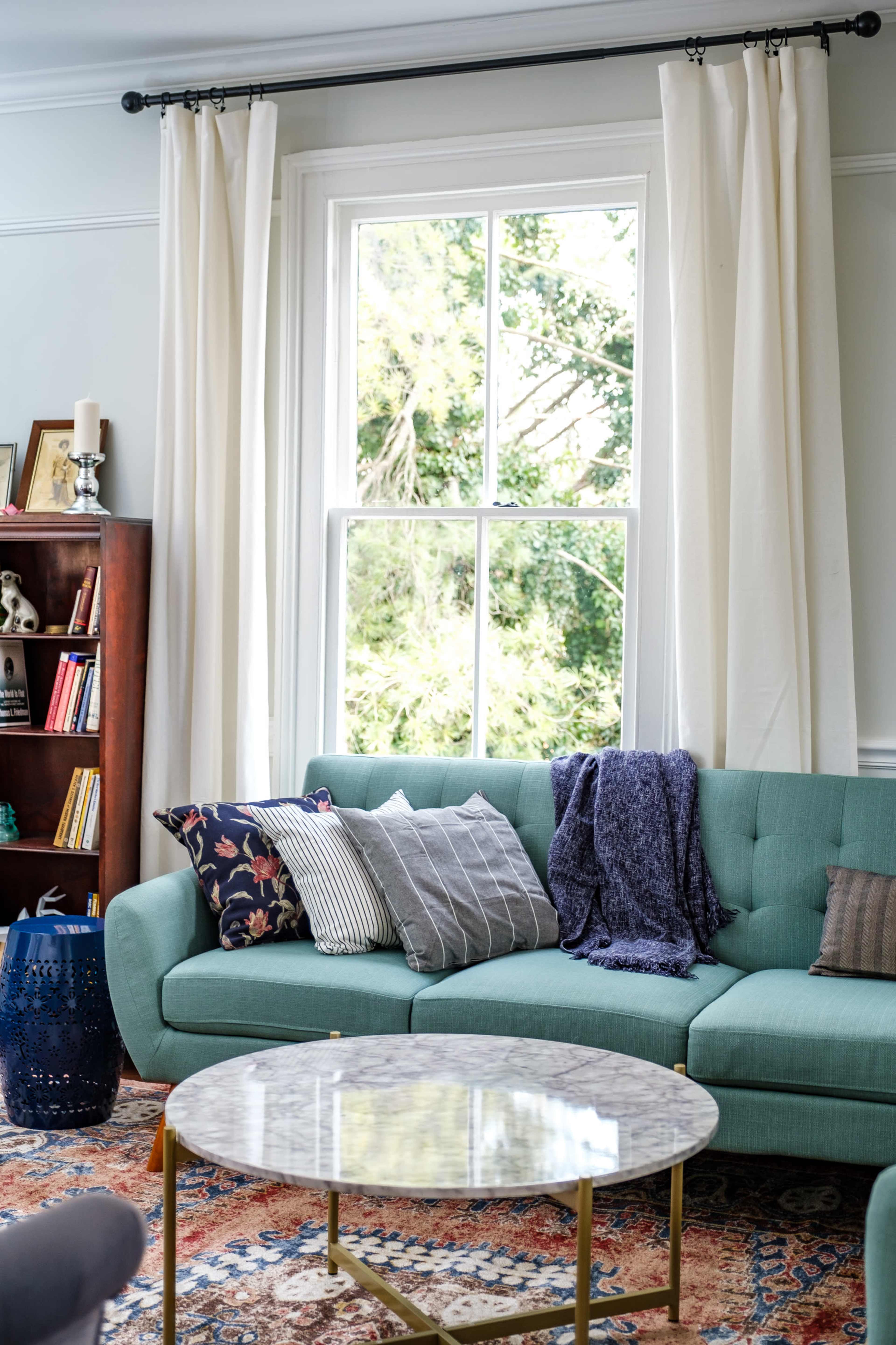 A light-filled living room with a teal sofa, a round marble coffee table, and bright curtains framing a window with a view of greenery outside.