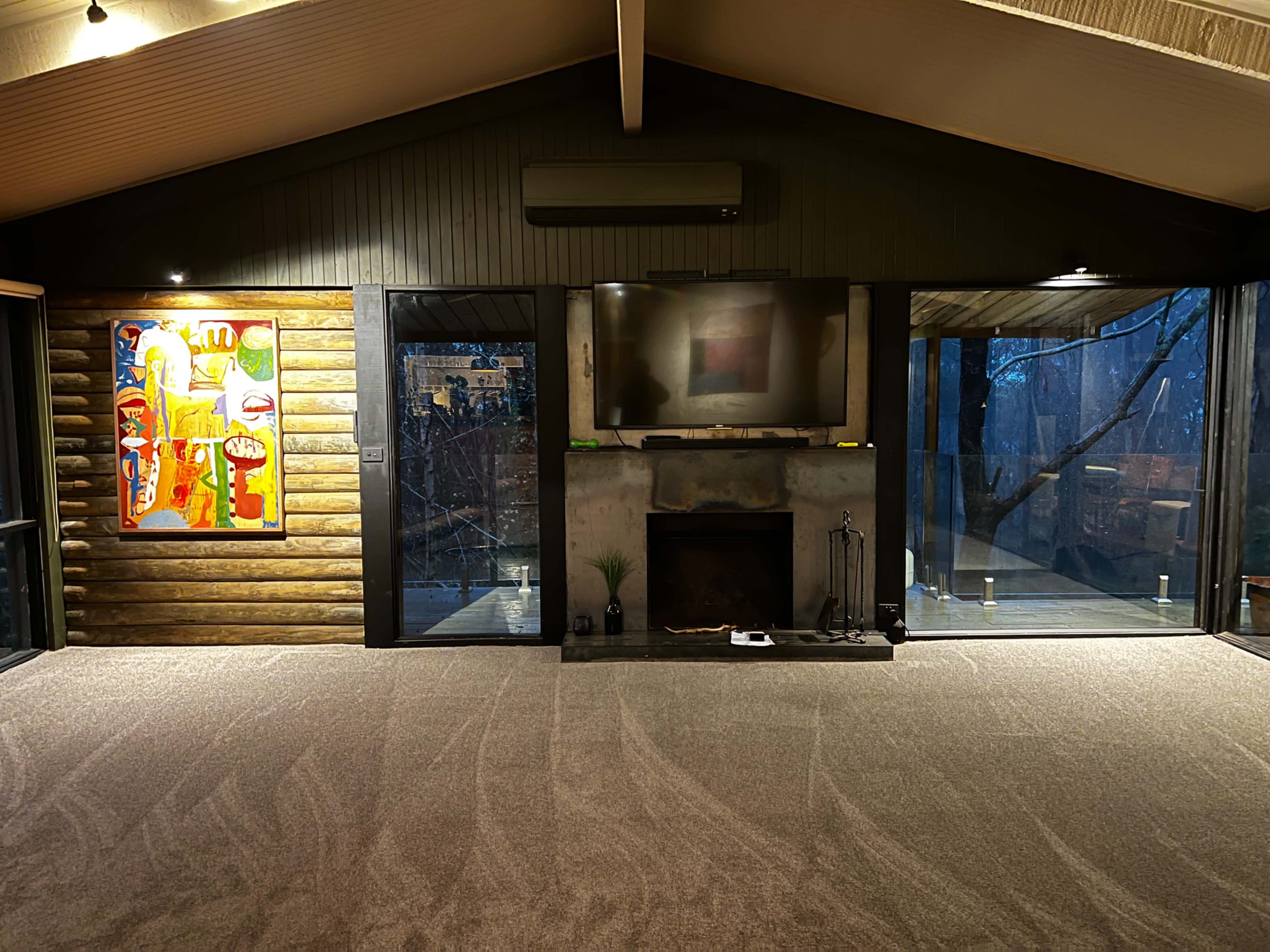 A modern room with wooden walls, a large television above a fireplace, and a colorful painting on the left, all illuminated by soft lighting.