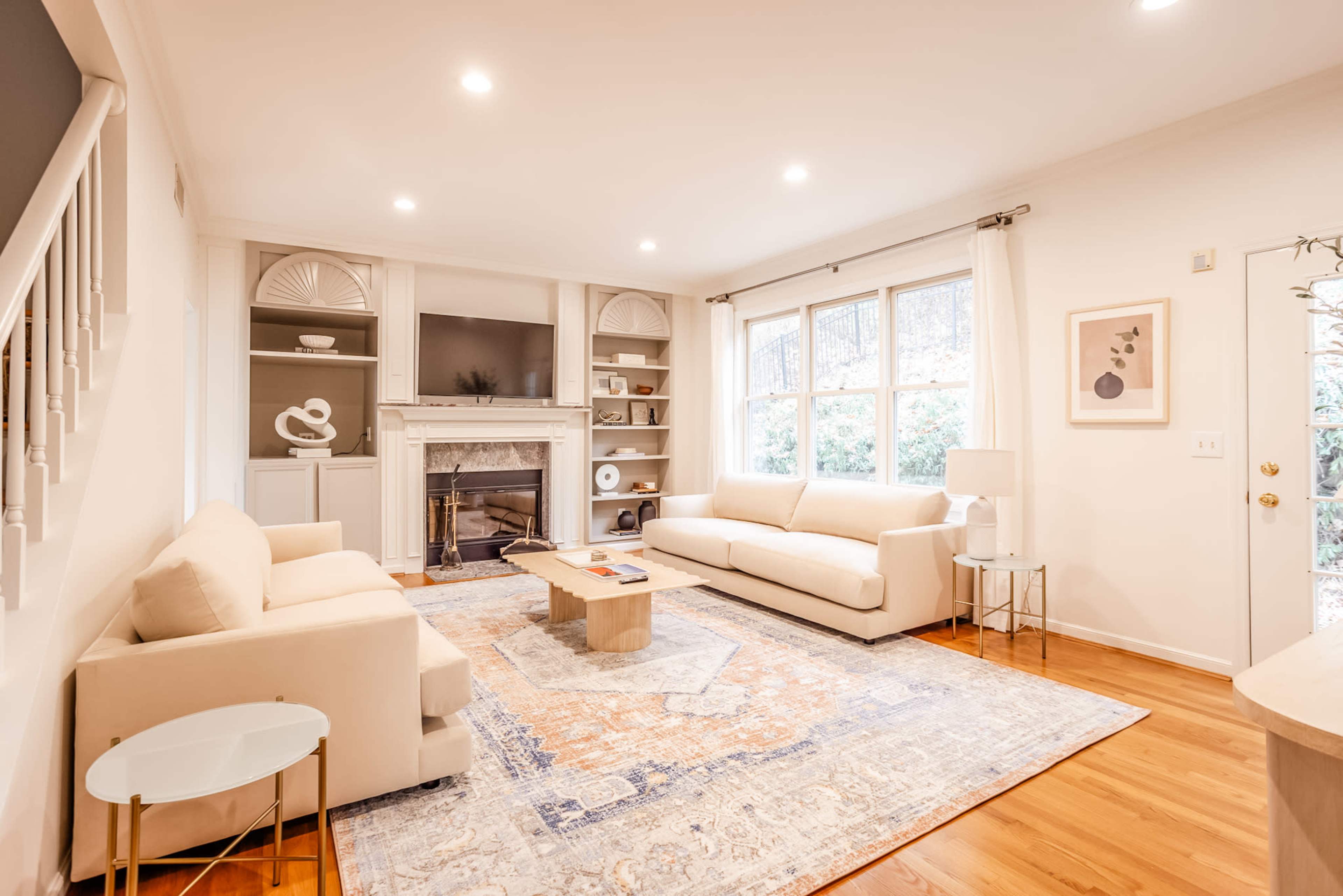 A bright living room features two beige sofas, a wooden coffee table, and a fireplace, with a large area rug and windows allowing natural light to enter.