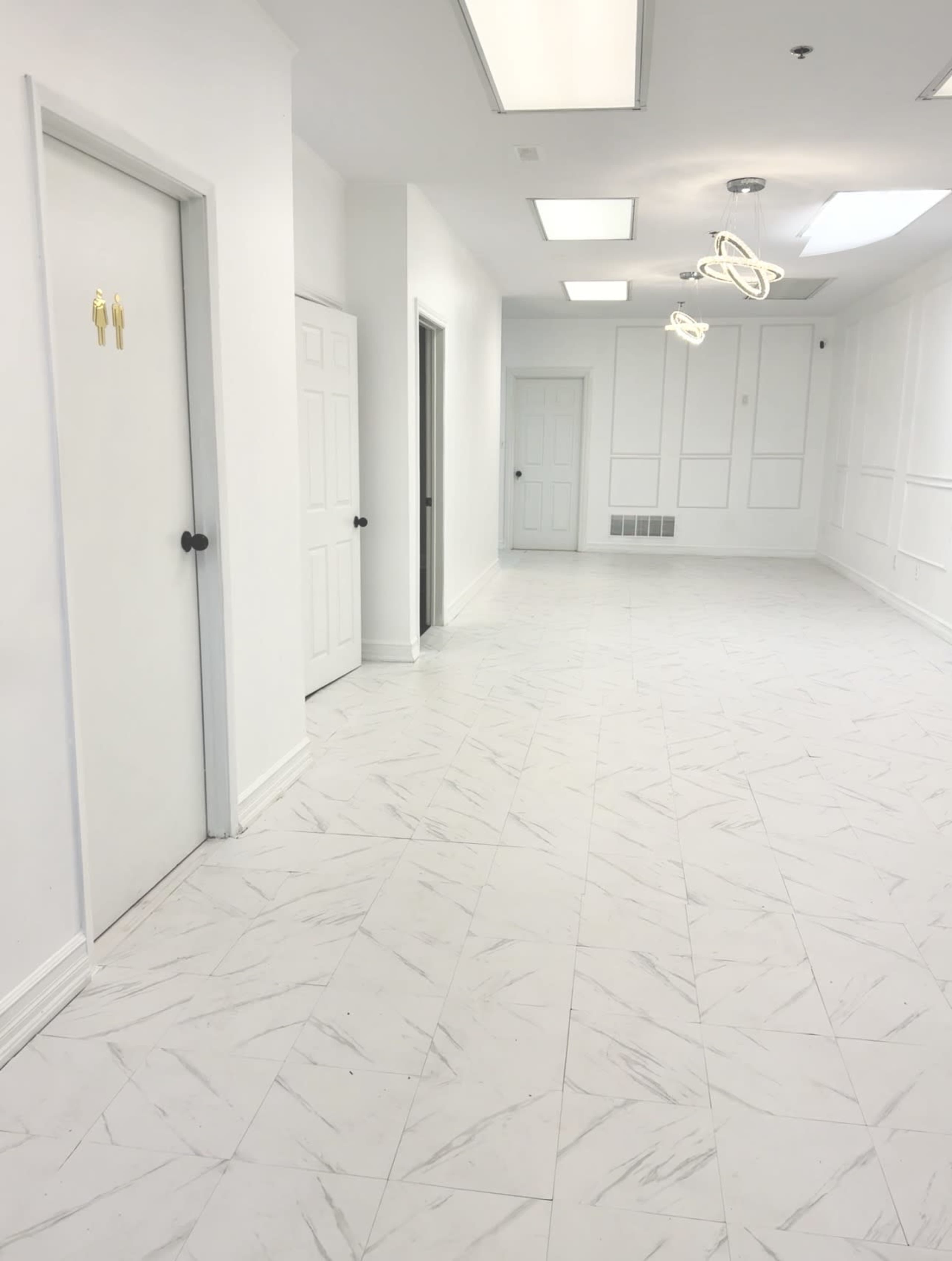 The image shows a spacious, bright corridor featuring several closed doors and a modern light fixture hanging from the ceiling.