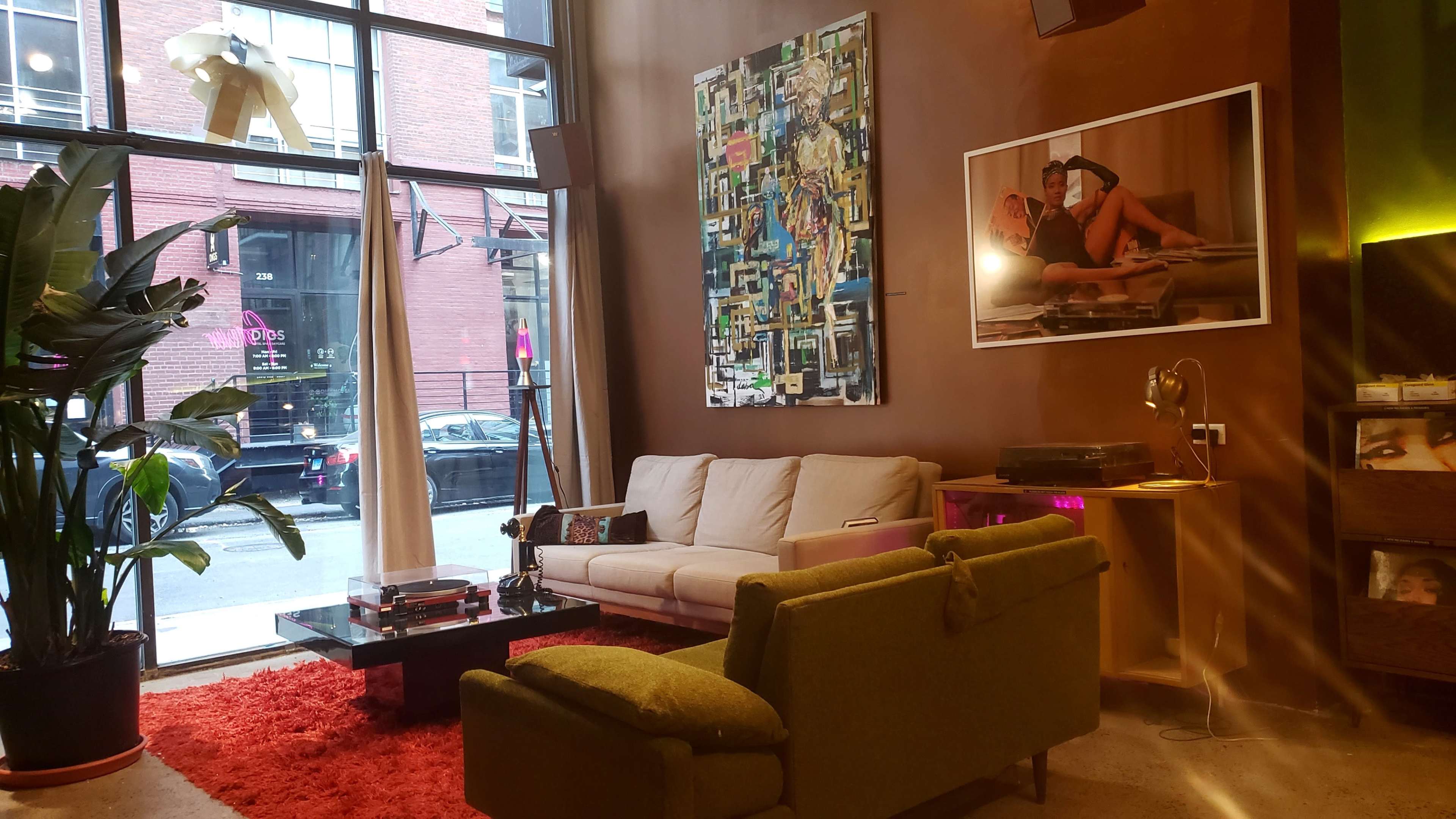 The interior of a modern lounge features a white couch and green armchair facing a coffee table, with large windows showcasing an urban street view and artwork on the walls.