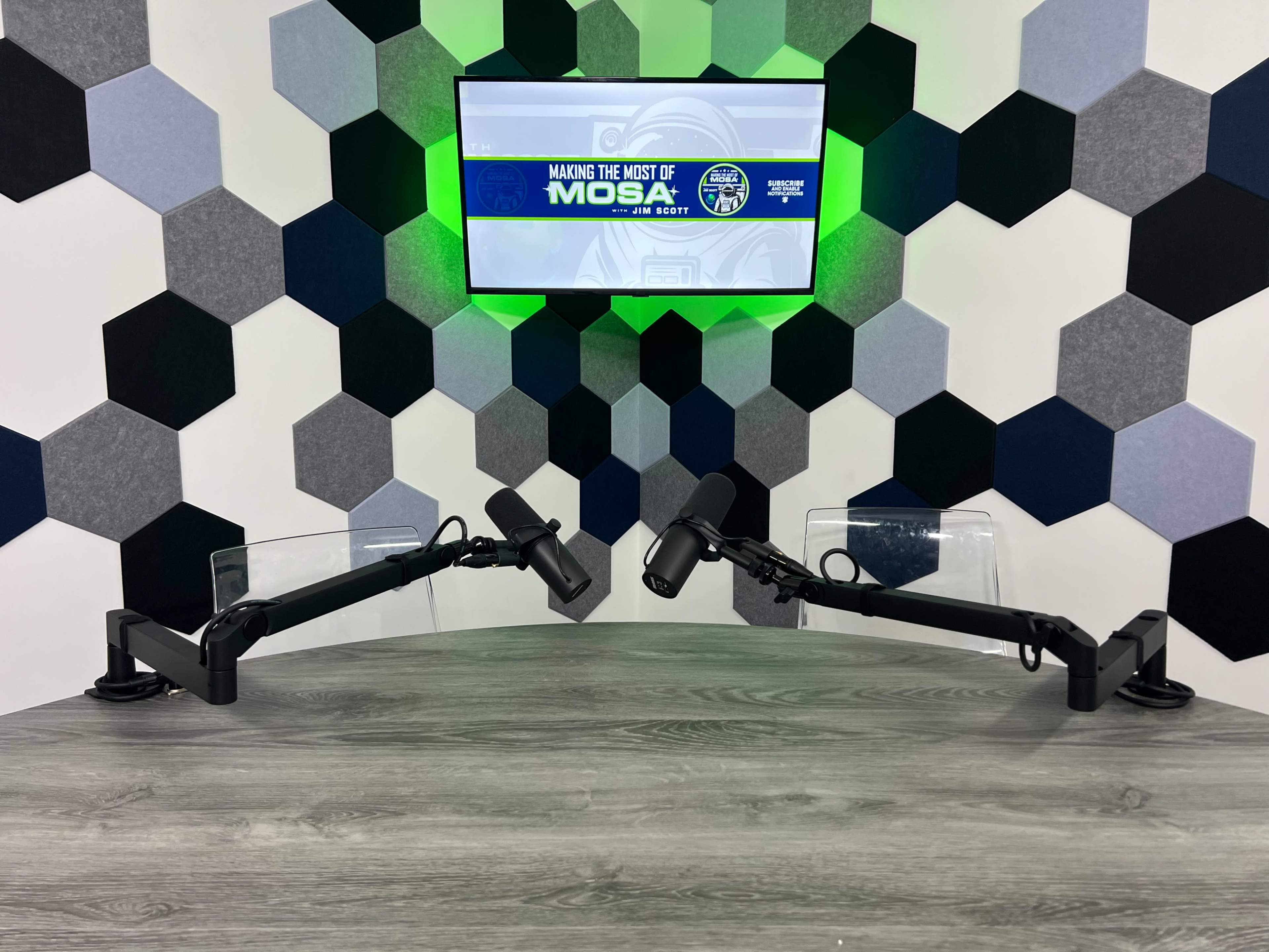 A modern podcasting setup features two microphone arms positioned on a wooden table, with a hexagonal-patterned wall backdrop and a screen displaying "Making the Most of Mosa."