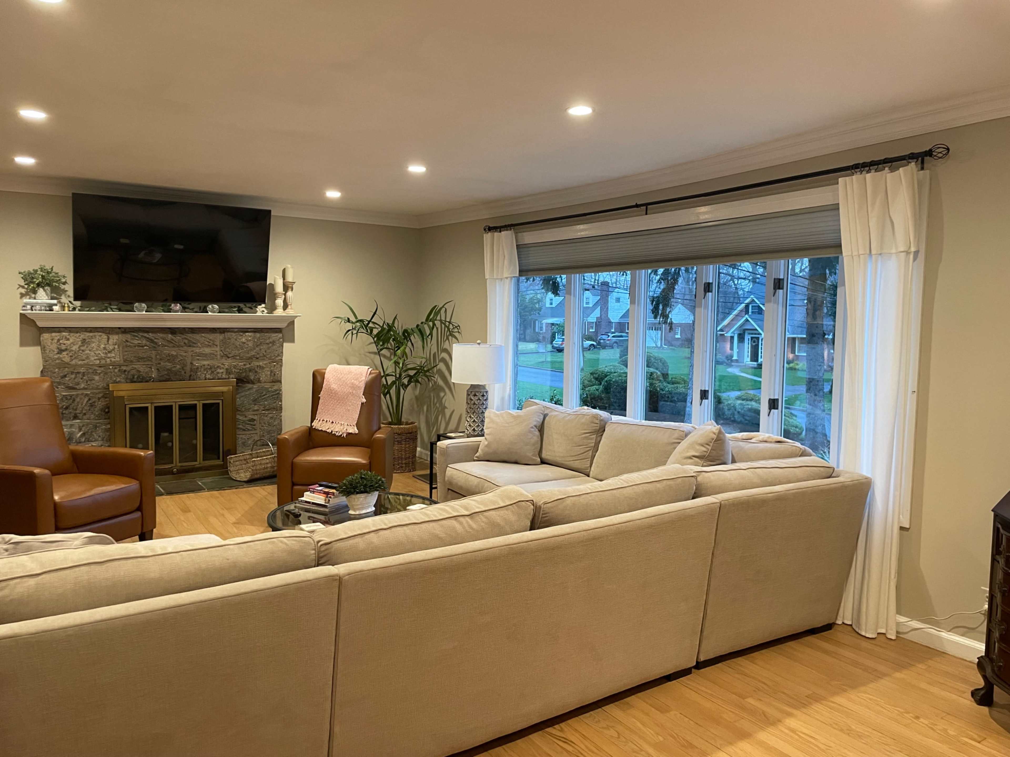 The living room features a cozy arrangement of beige sofas, two brown armchairs, a stone fireplace, and a large window with a view of the front yard.