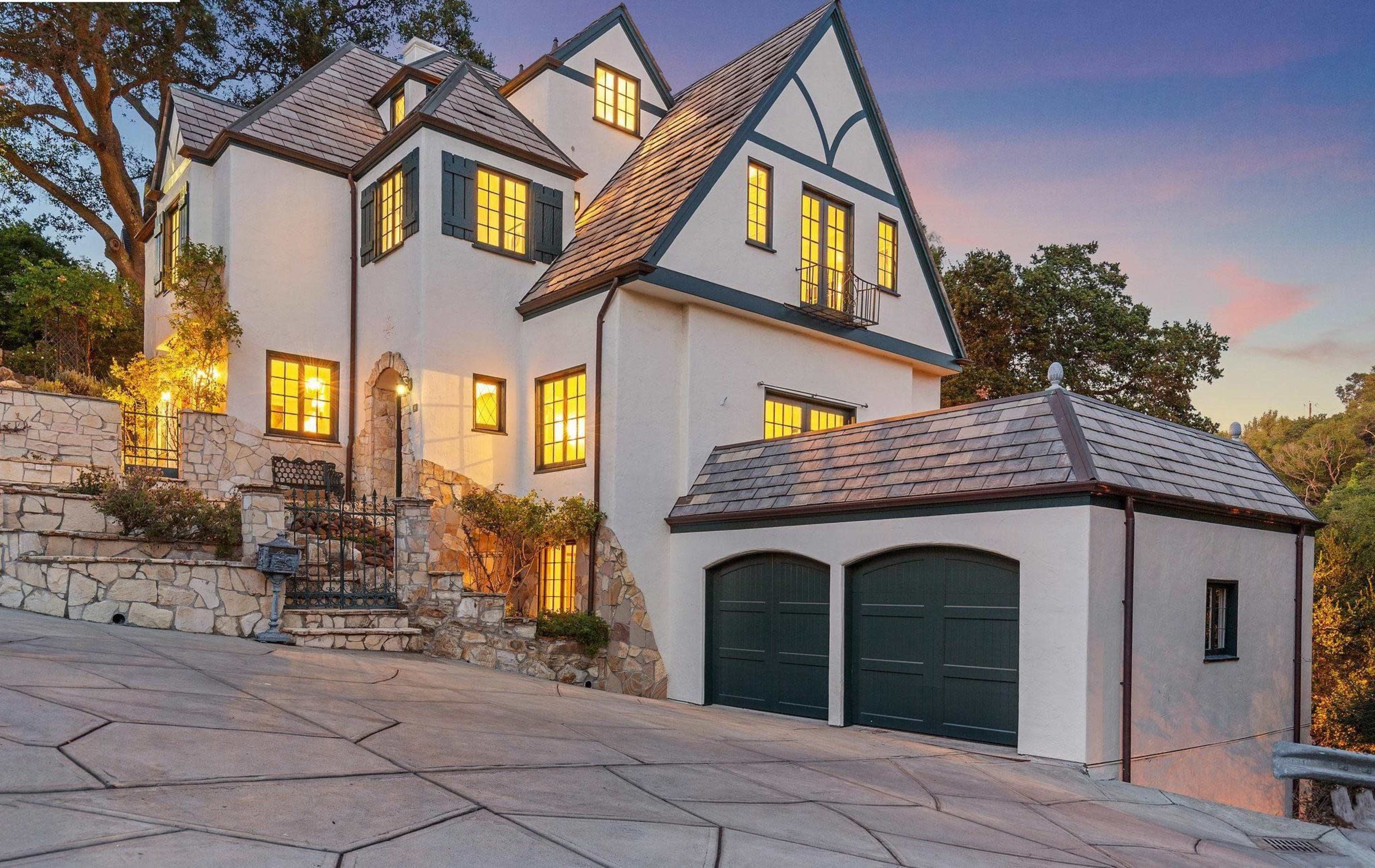 A multi-story house with a steep roof, large windows, and a stone pathway is situated on a sloping driveway surrounded by trees.