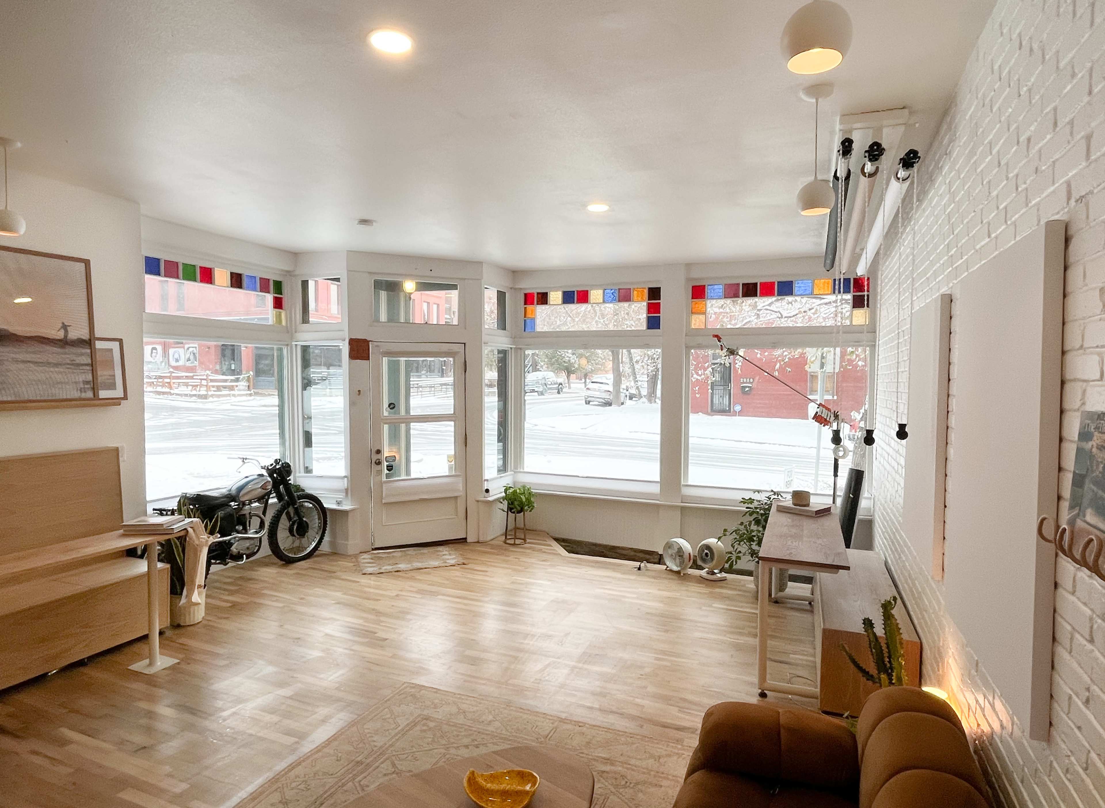 A bright, modern interior with large windows featuring colorful trim, a brown sofa, a wooden table, and a motorcycle parked by the entrance.