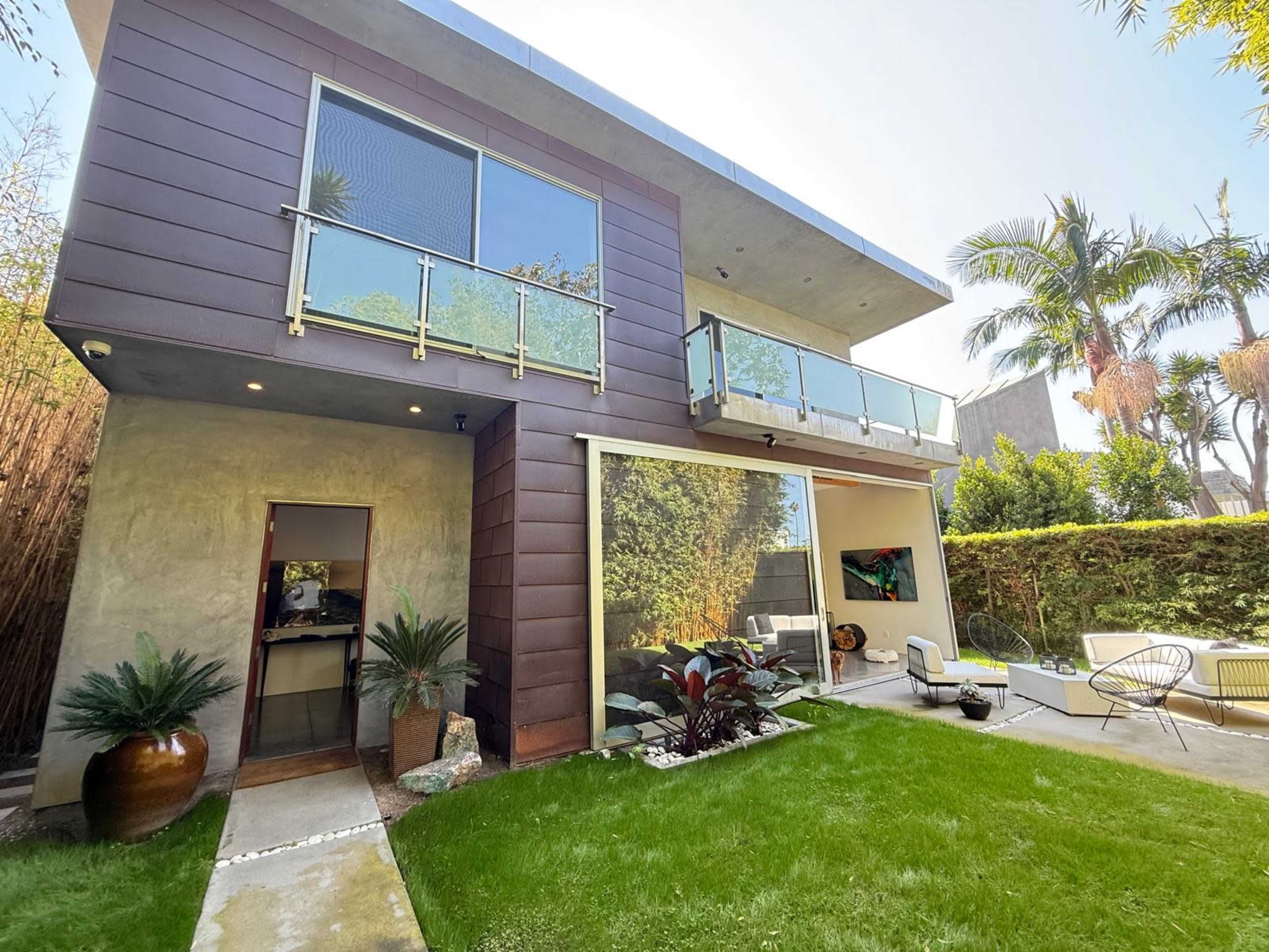 The image shows a modern two-story house with large glass windows, a balcony, and a well-maintained garden area.