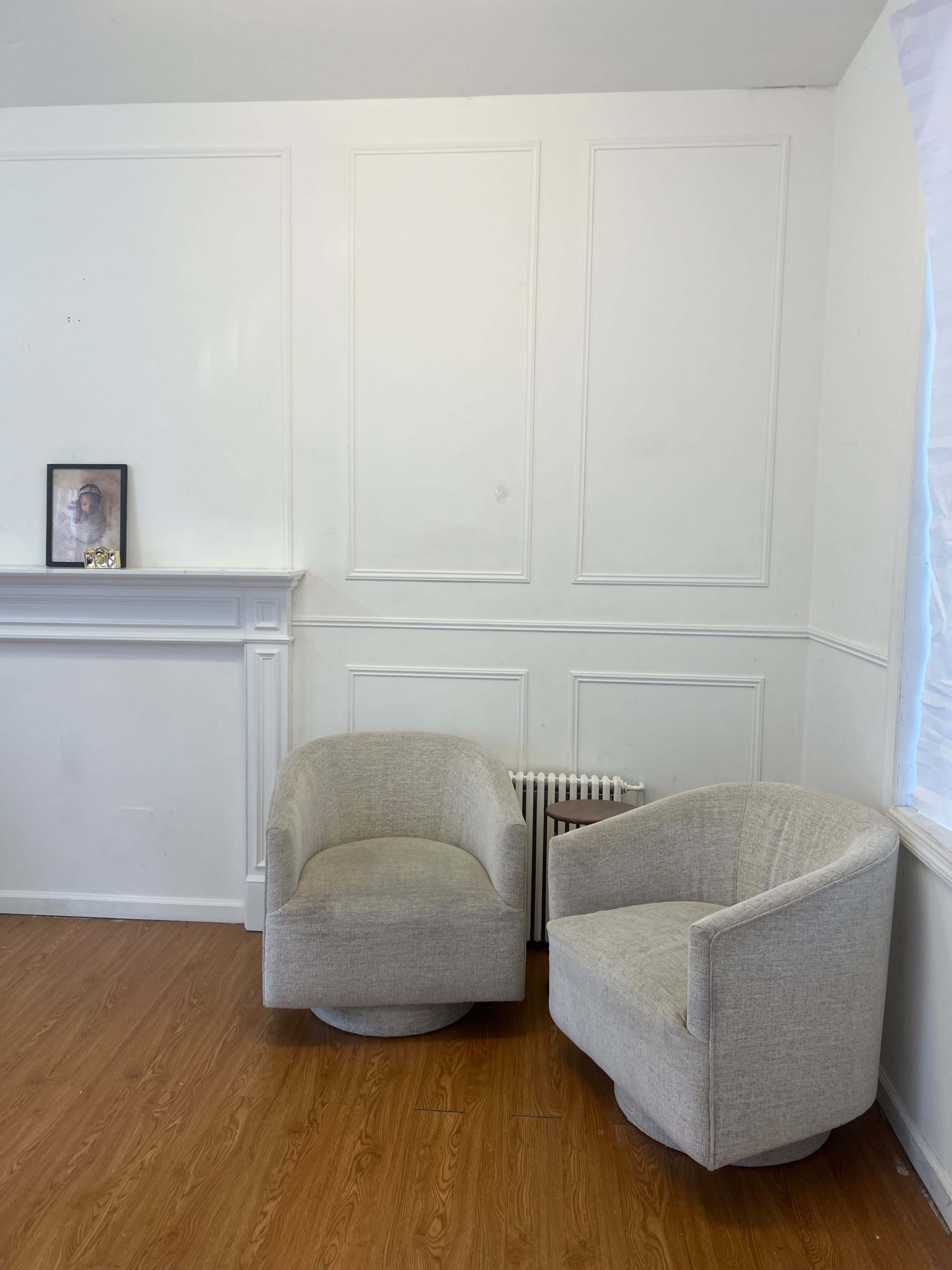 The image shows a minimalist room with two light-colored lounge chairs beside a small side table and a wall-mounted picture.