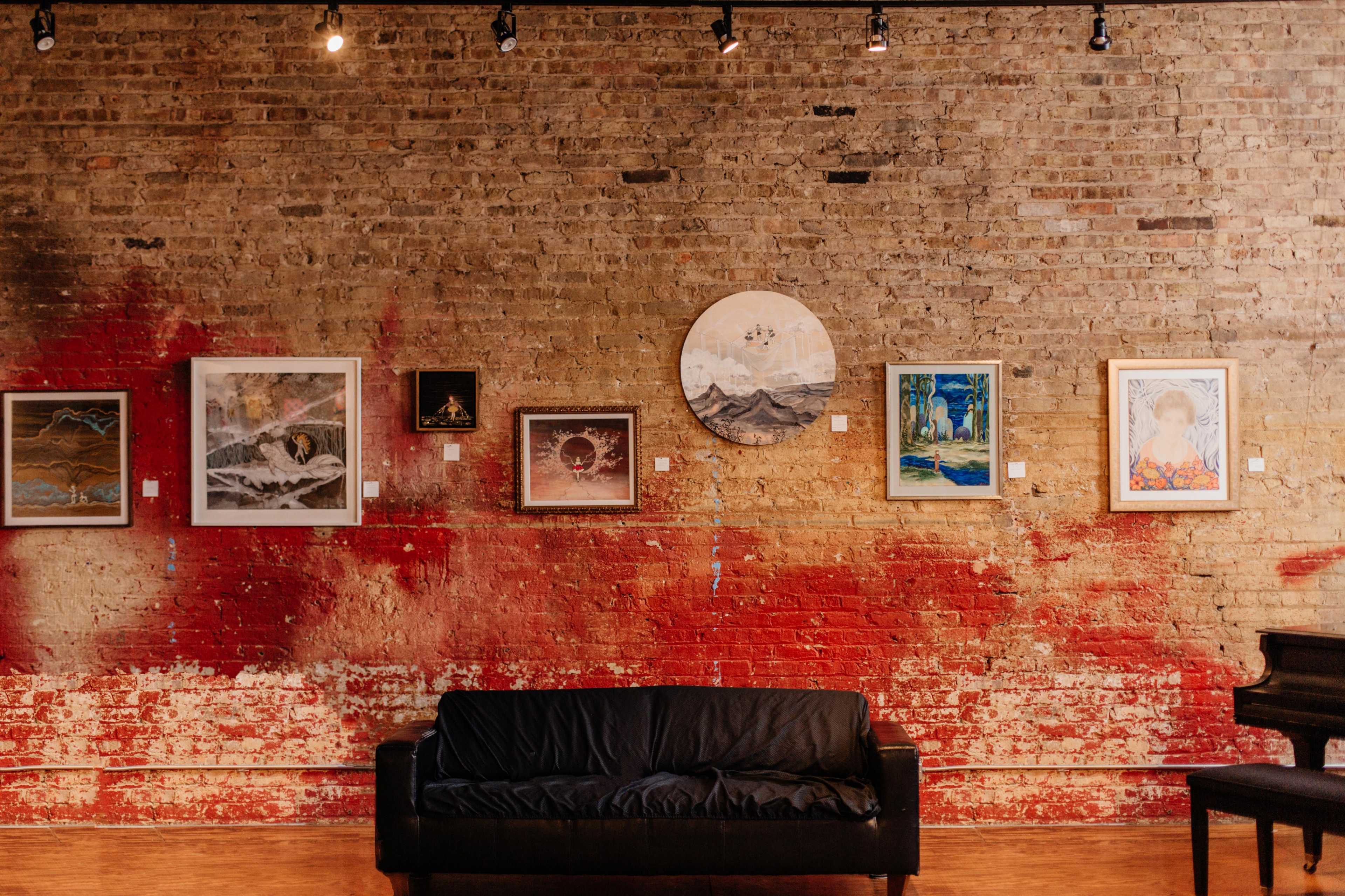 A gallery wall featuring various framed artworks displayed against a brick wall with a splash of red paint, alongside a black couch in the foreground.