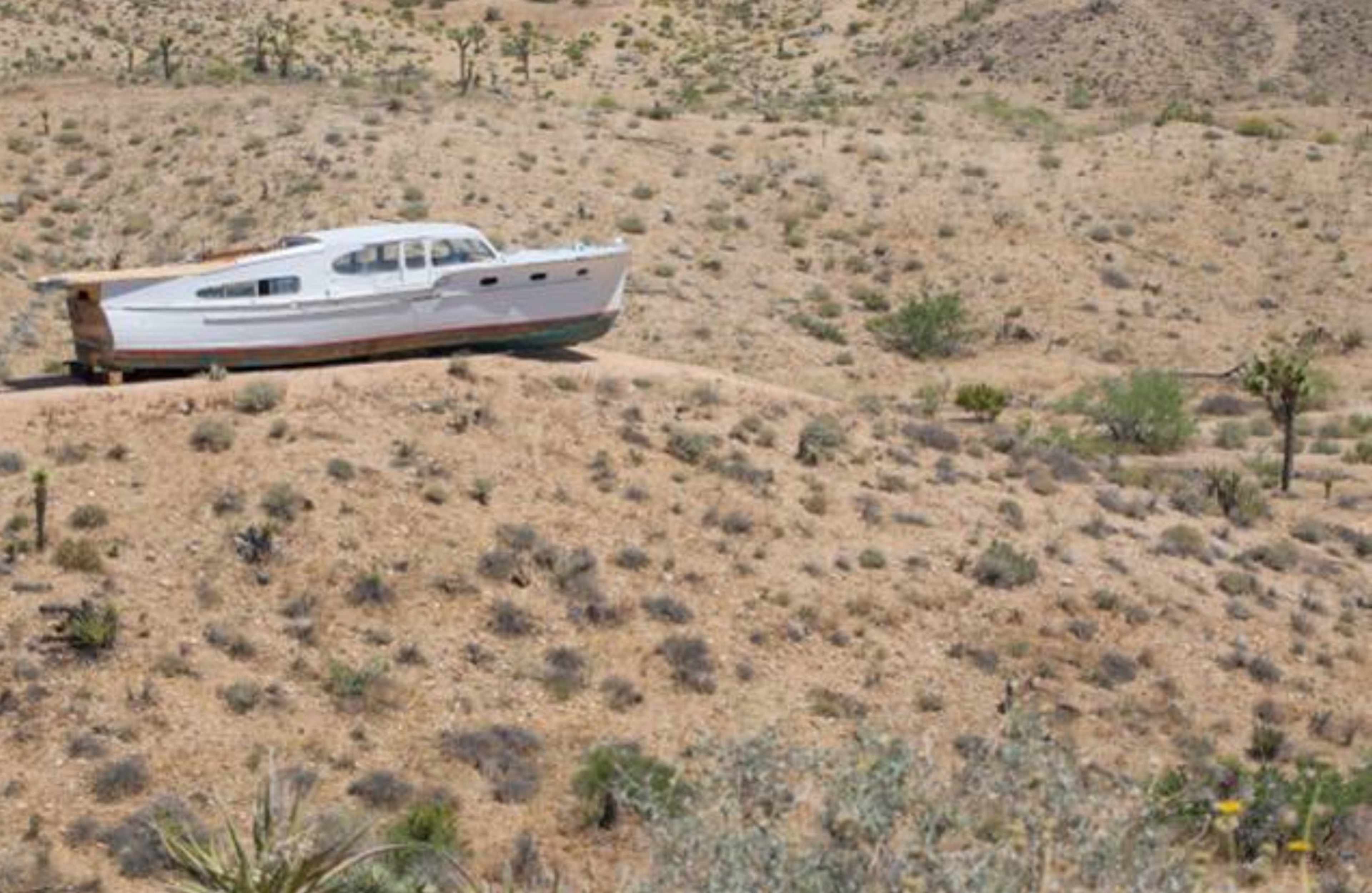 A white boat sits stranded on a dry, sandy hillside surrounded by sparse vegetation.