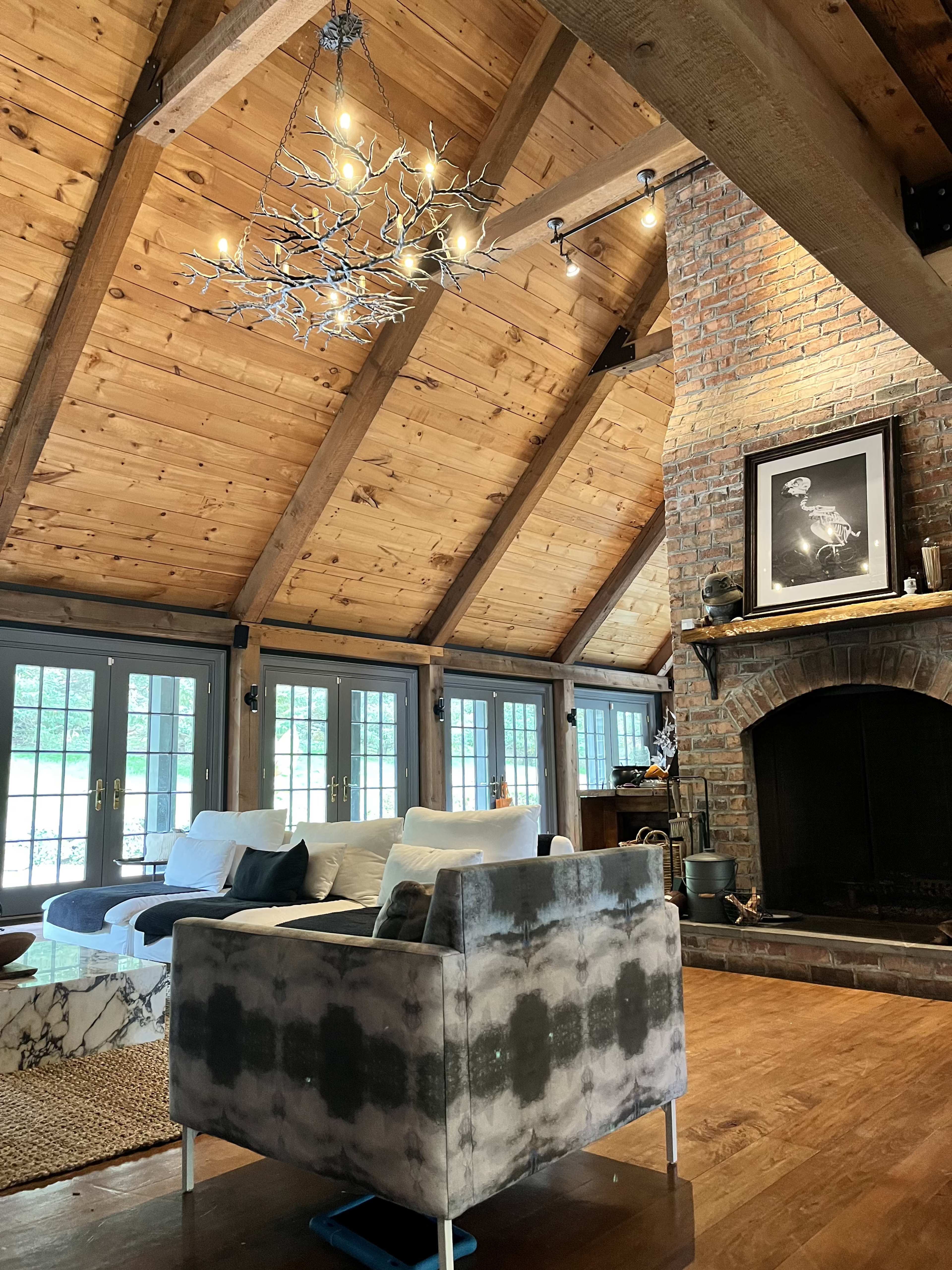 A cozy living room with a high wooden ceiling, a brick fireplace, and large windows.