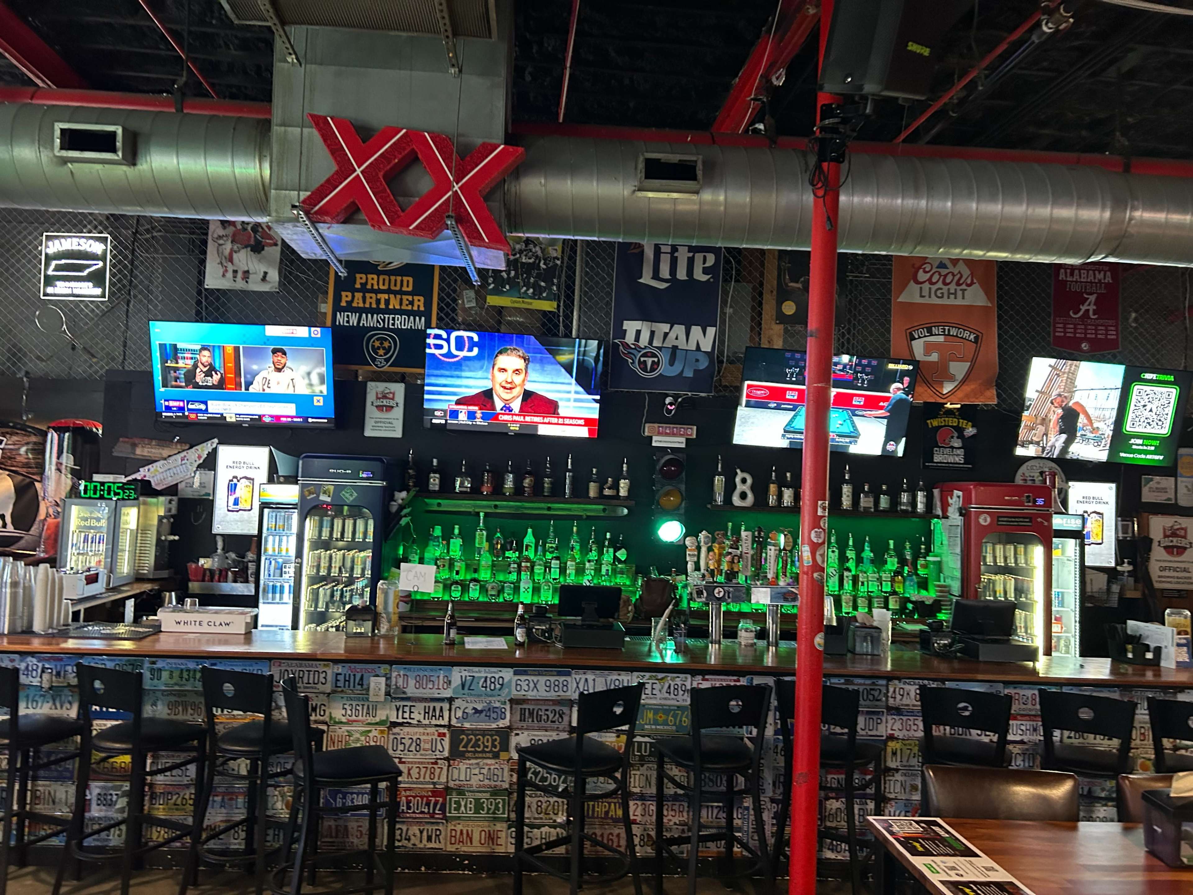 The image shows the interior of a sports bar with multiple televisions displaying sports events and various bottles of liquor illuminated behind the bar.