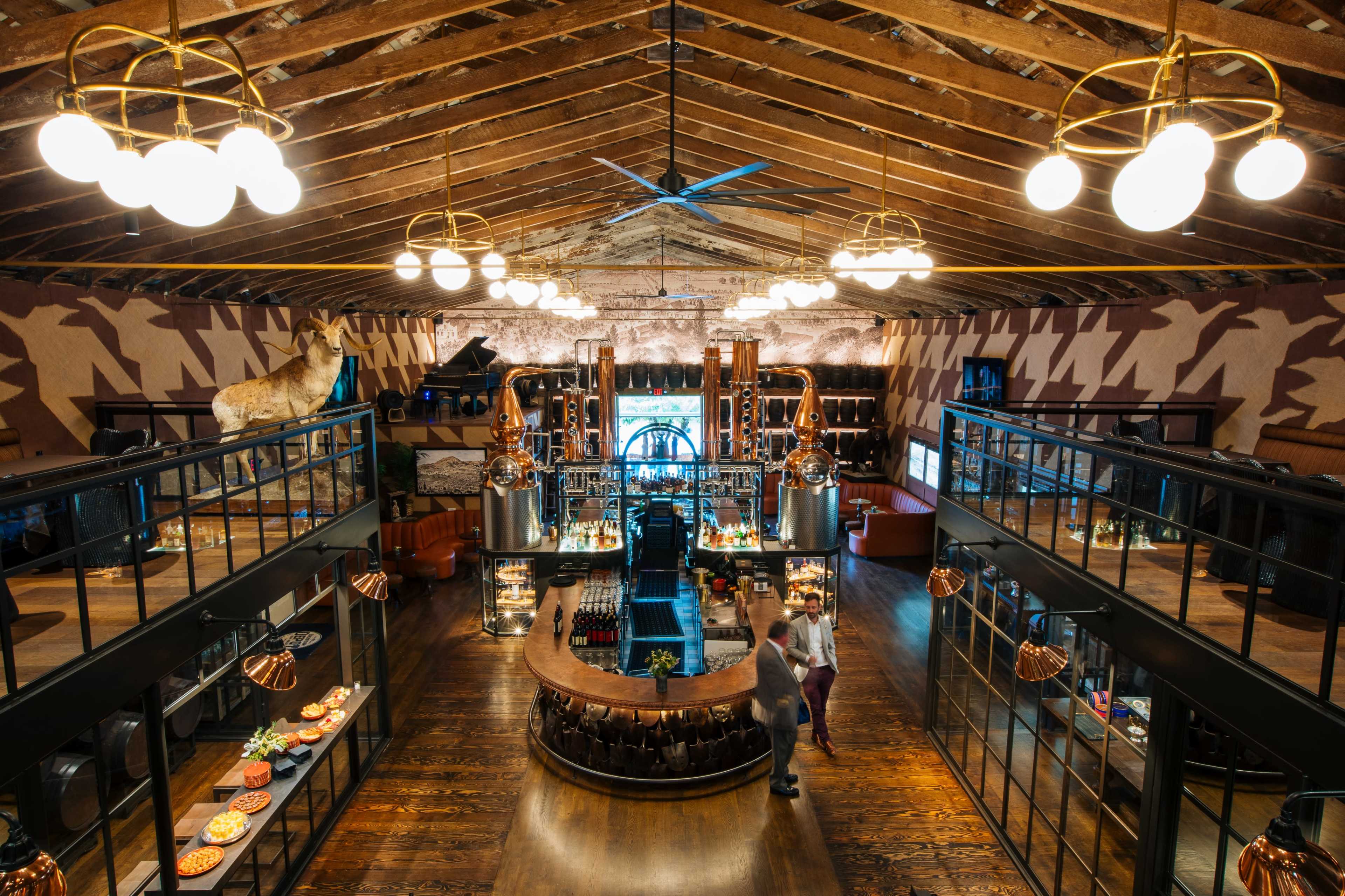 The image shows a spacious, warmly lit bar and dining area with high wooden ceilings, featuring a central bar surrounded by shelves of bottles and a variety of seating arrangements.