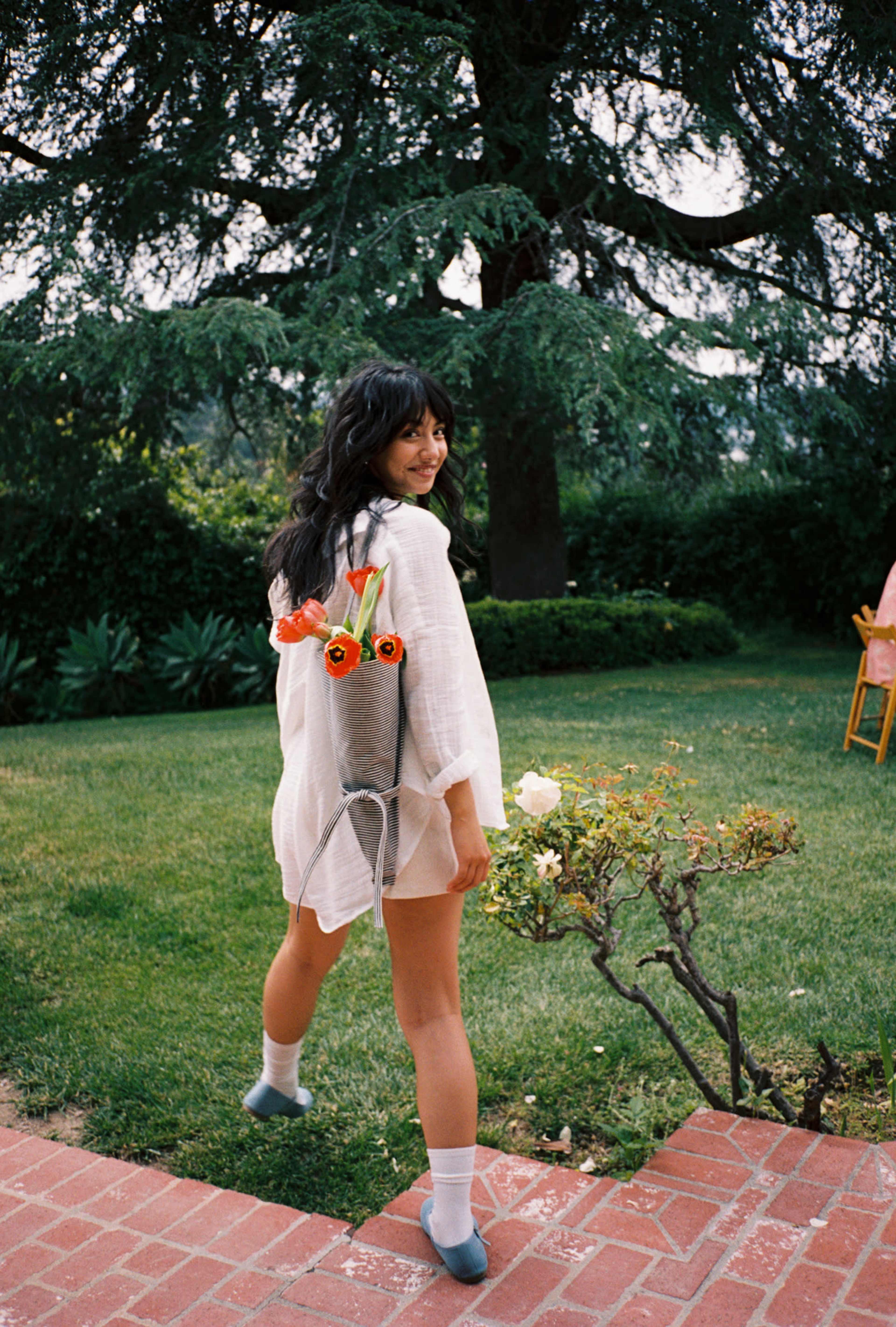 A woman walks away in a garden, wearing a light outfit with flowers attached to a bag on her back.