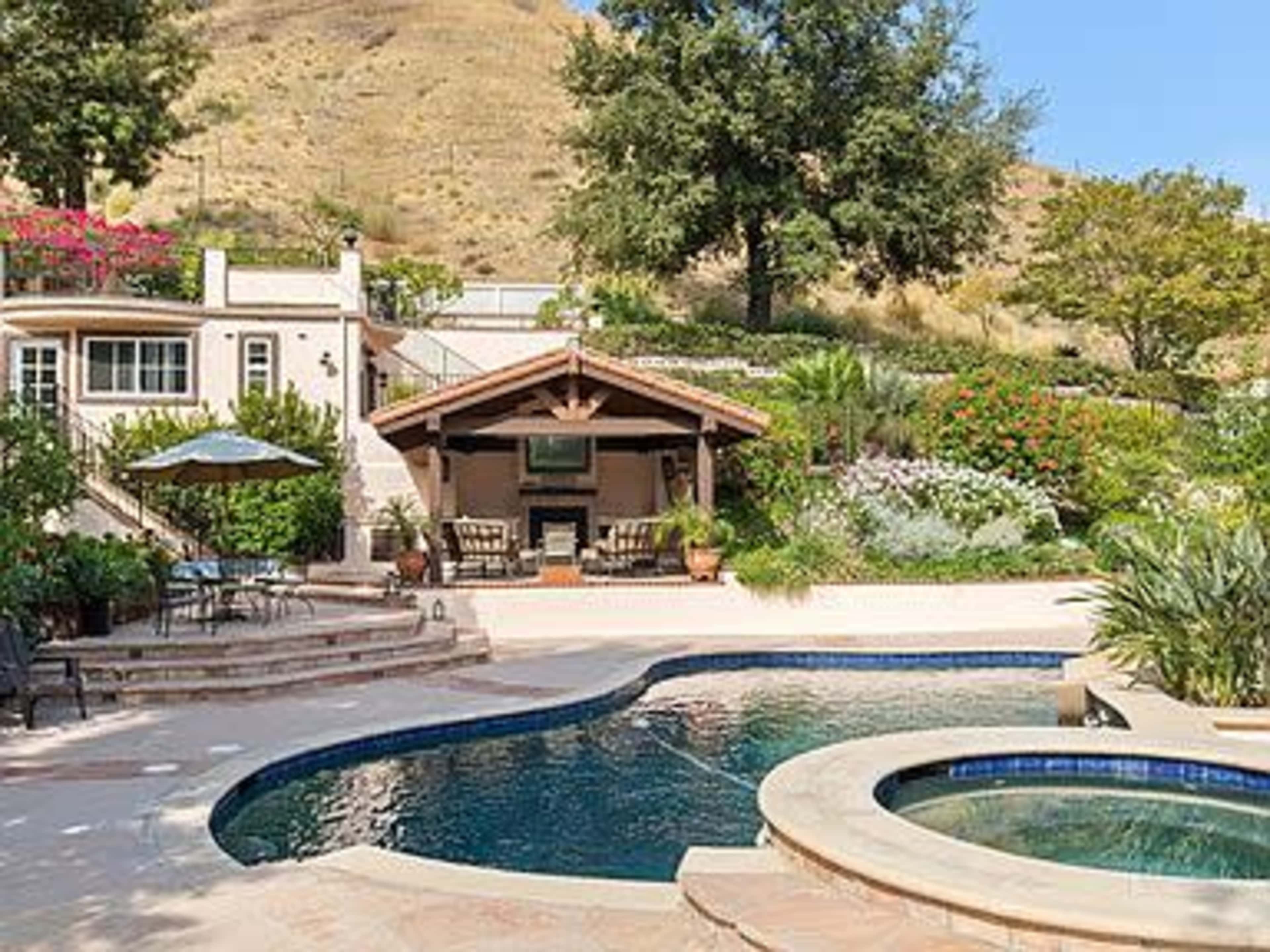 A landscaped backyard with a swimming pool, lounge chairs, and a patio adjacent to a house and hillside.