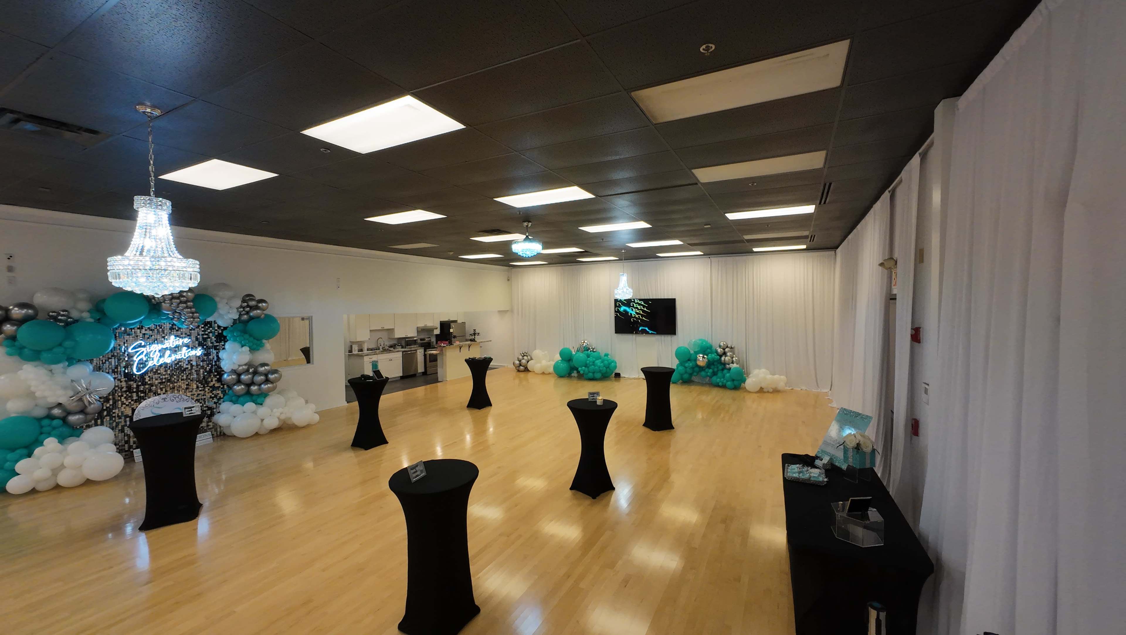 A spacious event venue decorated with black tables, teal and white balloons, and a chandelier.