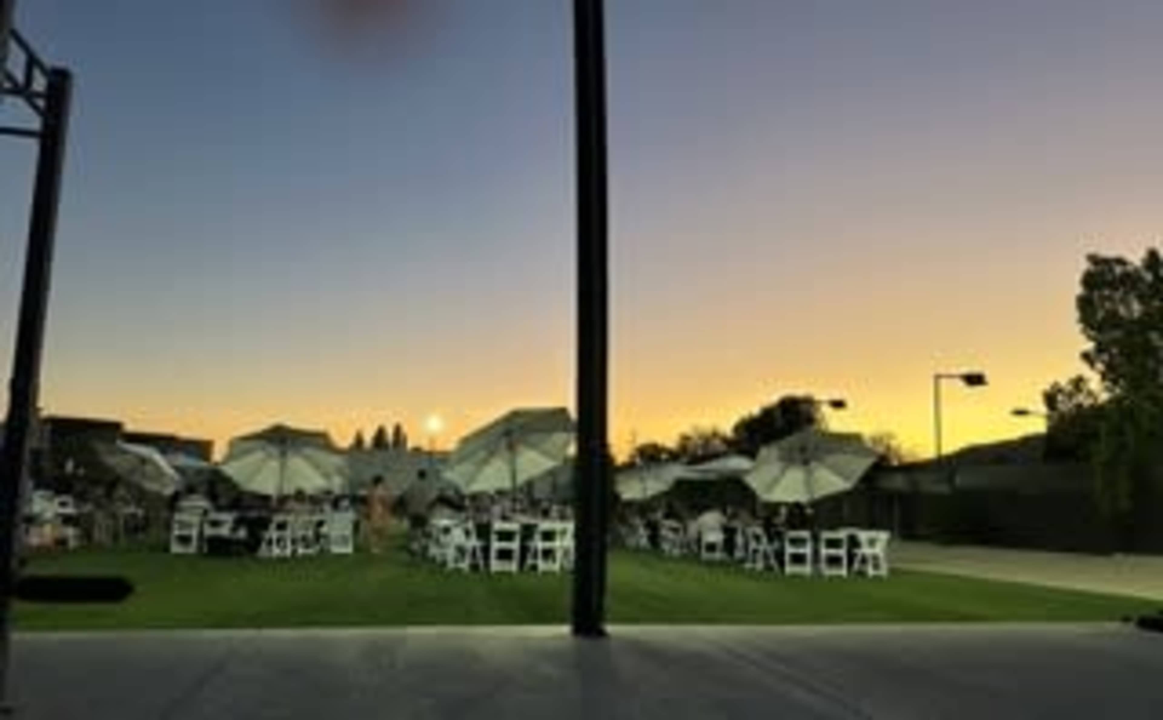 A set of white tables and chairs is arranged on a grassy area under large umbrellas, with a colorful sunset in the background.