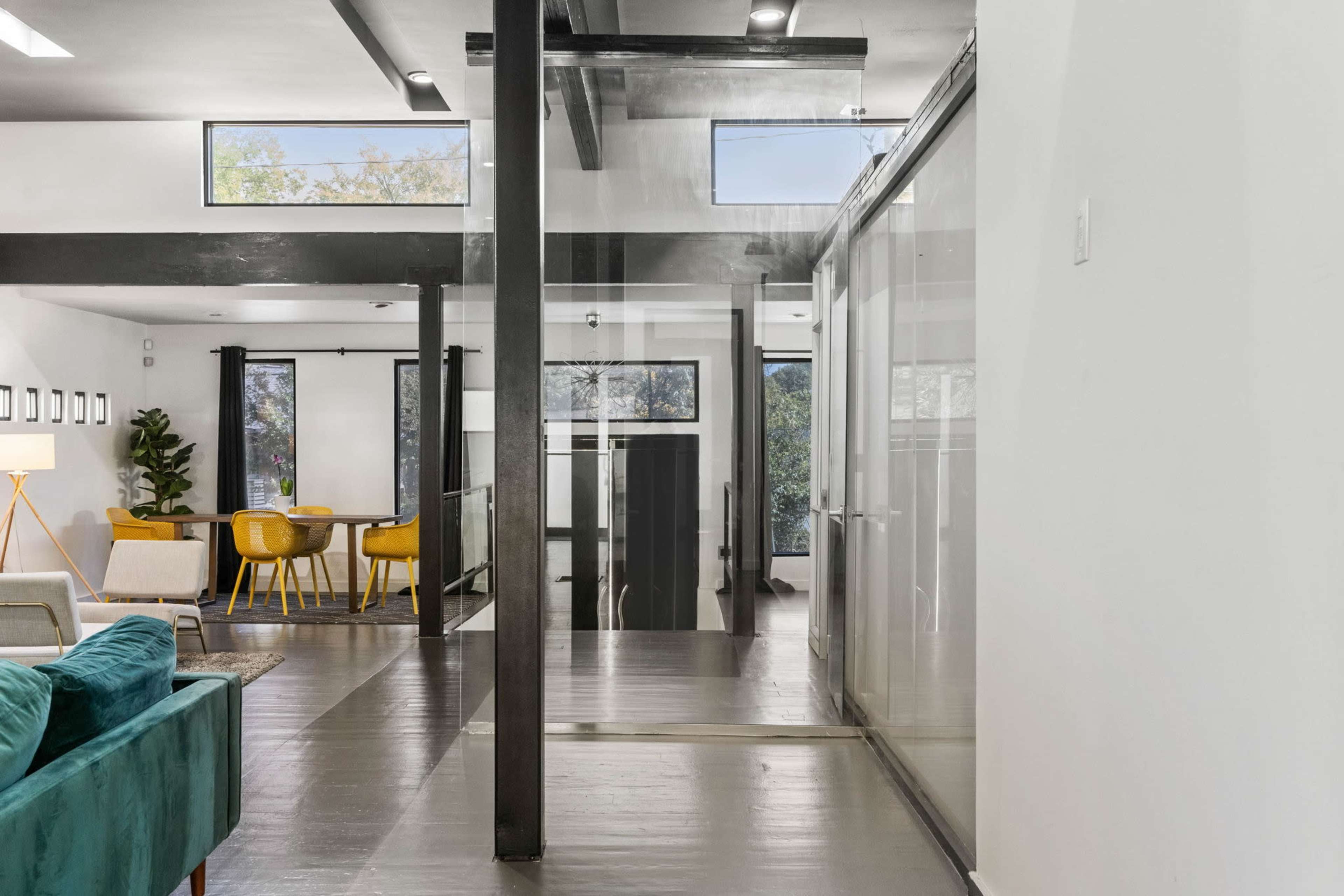 The image shows a modern interior space featuring a clear glass wall, open layout, and a living area with a green sofa and yellow dining chairs.