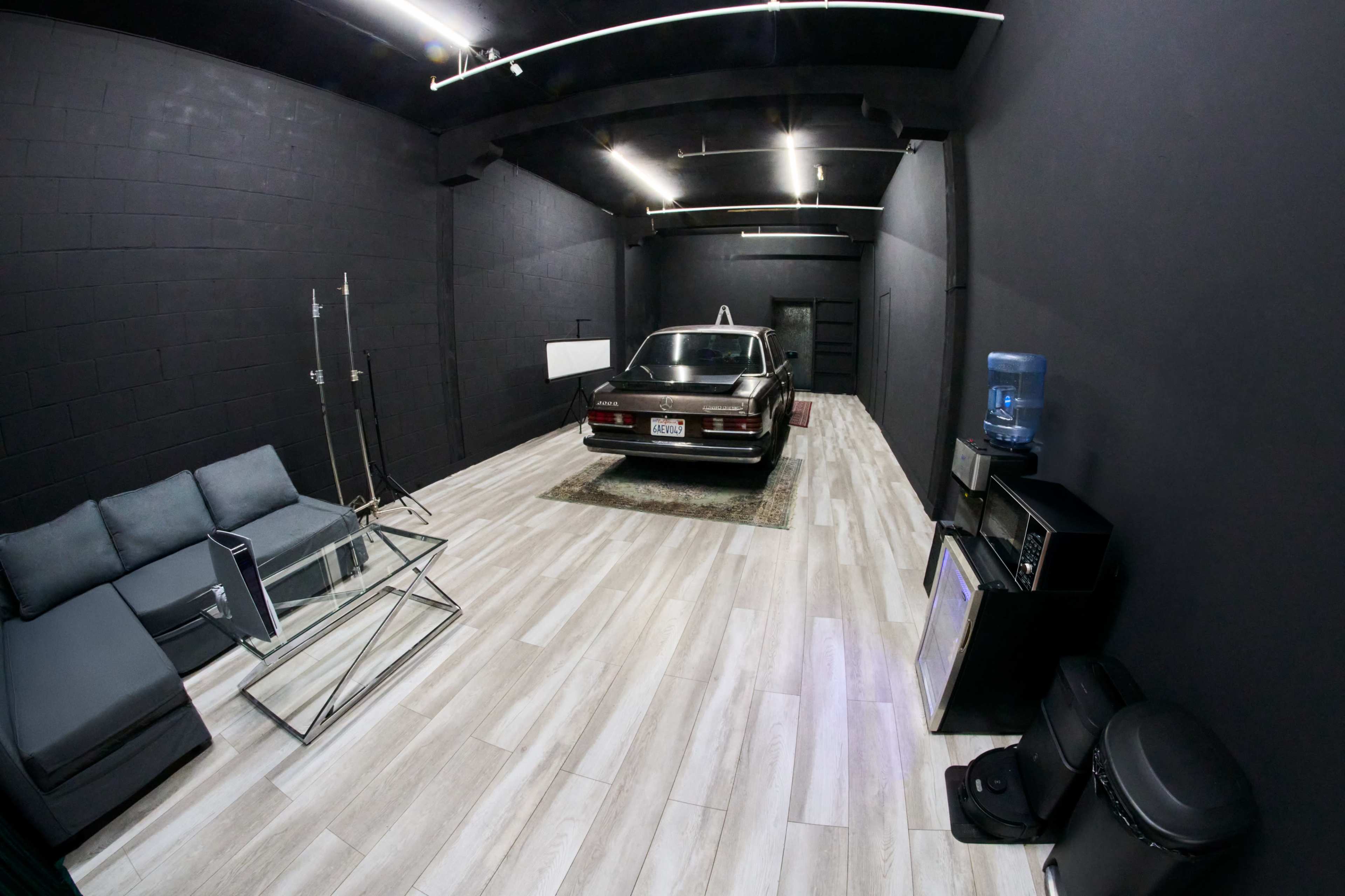 The image shows an empty room with dark walls, a vintage car parked in the center, and a seating area with a couch and glass coffee table.