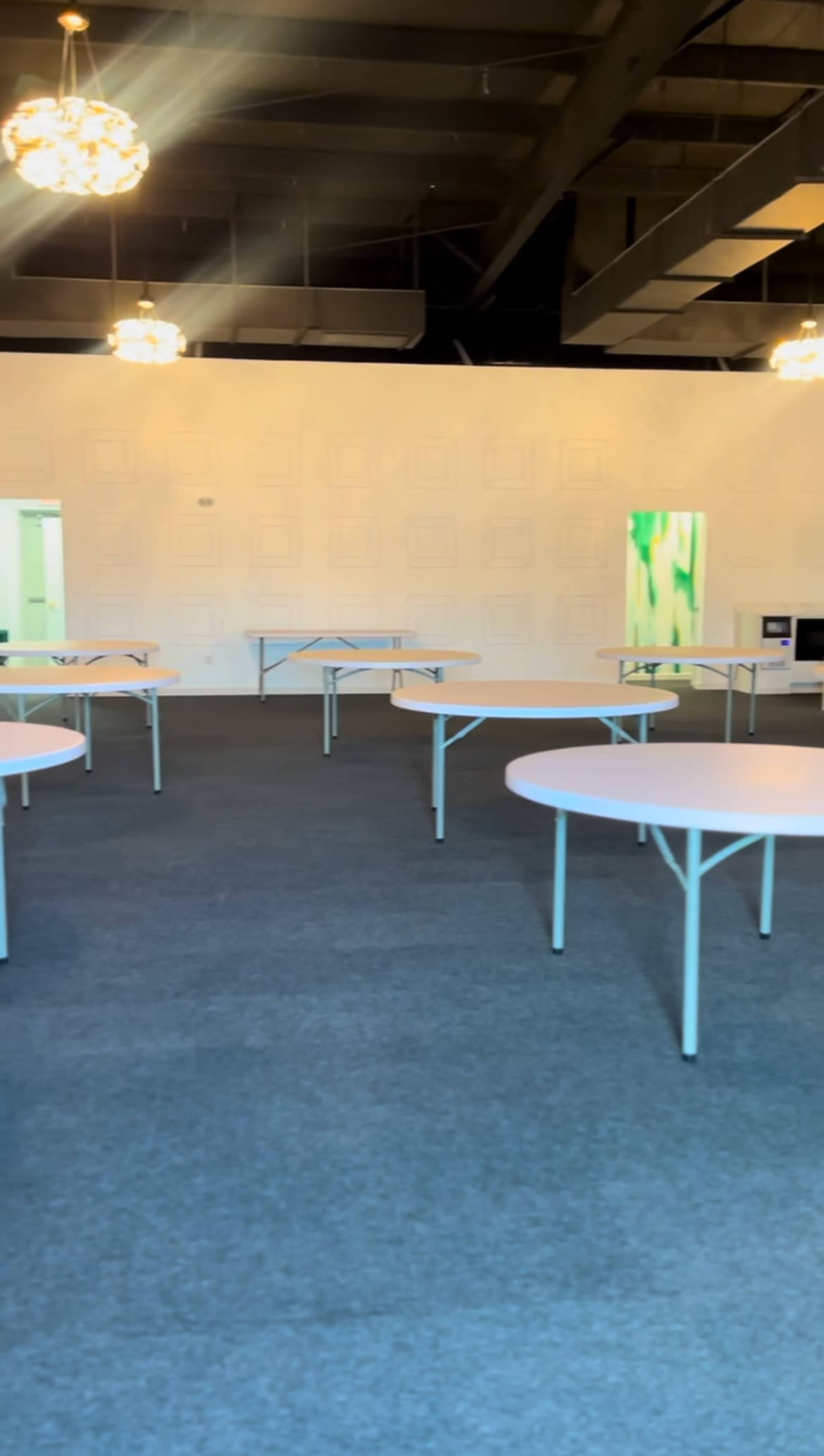 The image shows a spacious room with several white folding tables arranged evenly across a gray carpet.