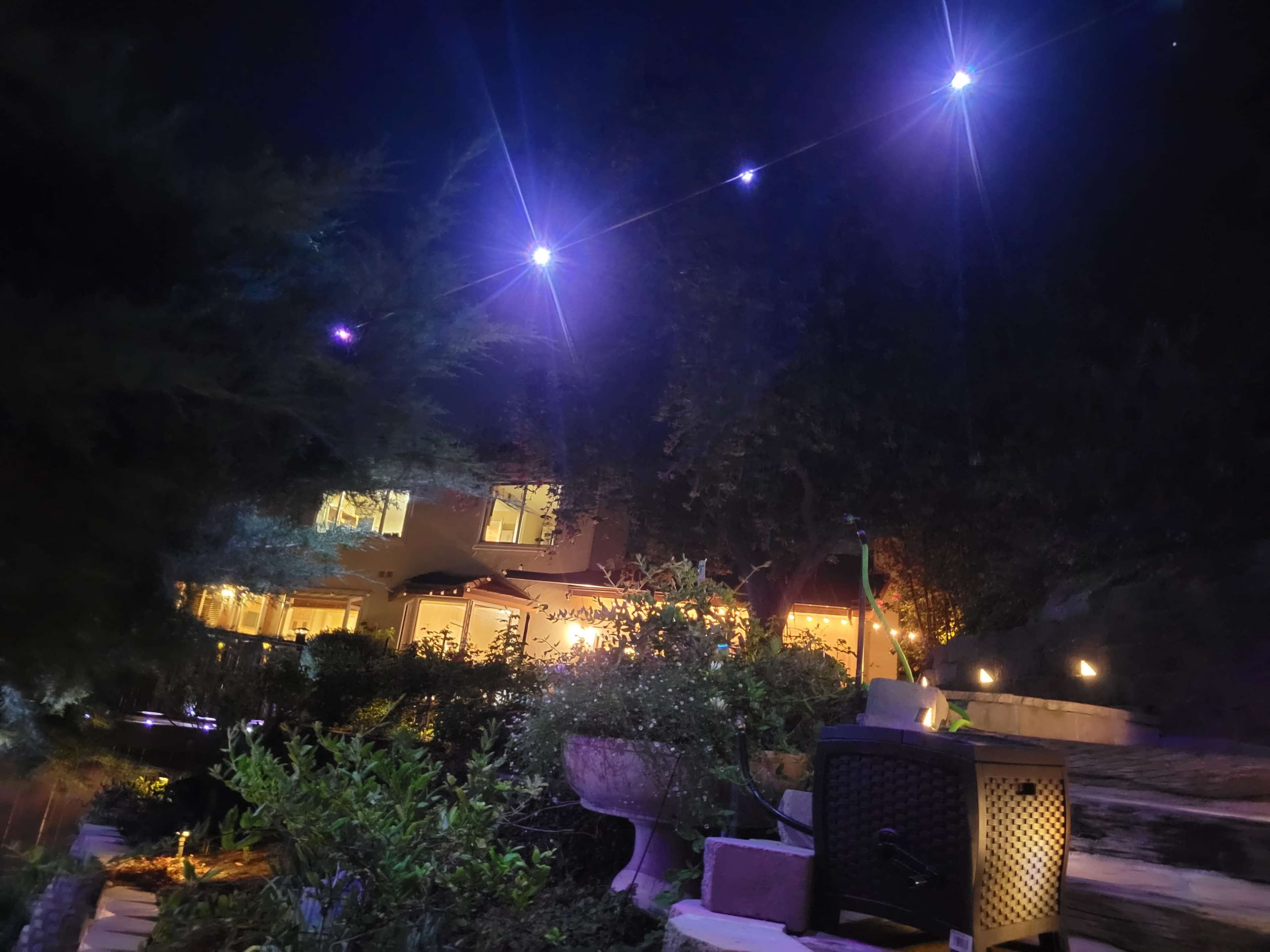 The image shows a well-lit garden area at night, featuring a house with illuminated windows and string lights overhead.