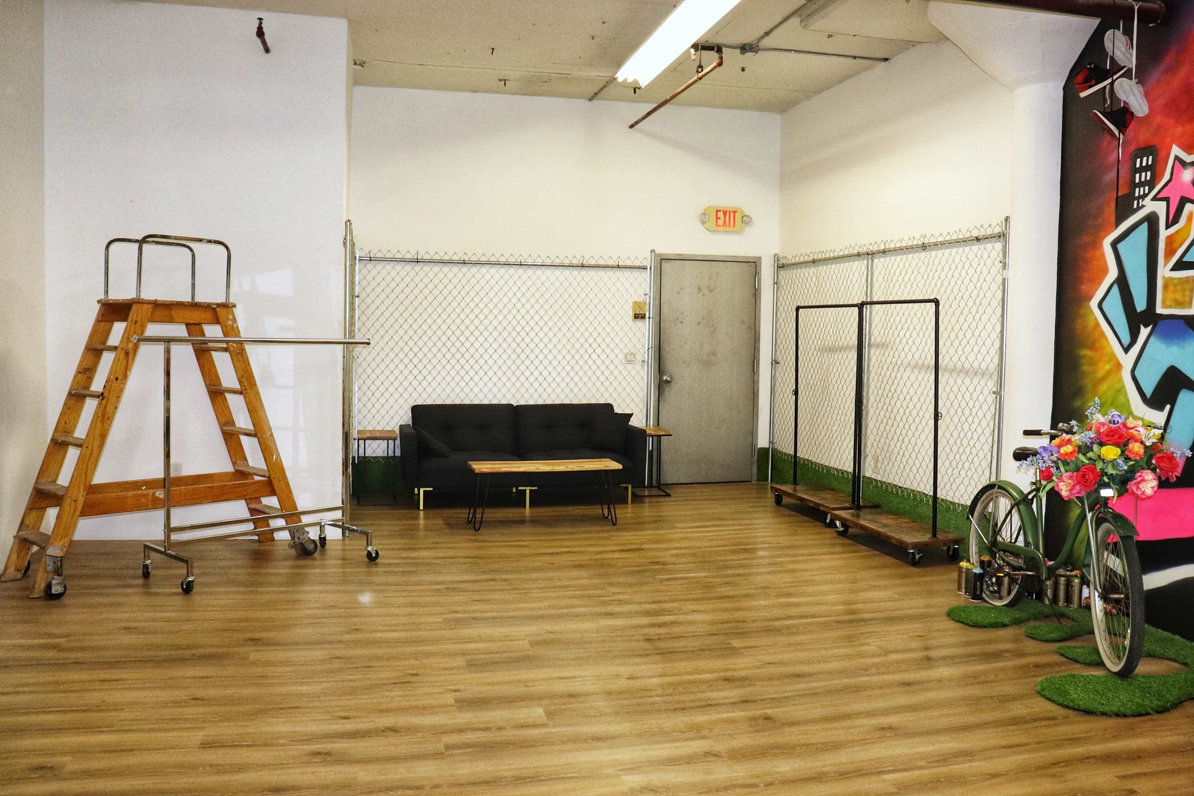 An empty interior space with a wooden ladder, a black couch, a table, a chain-link fence, and a bicycle next to decorative flowers against a colorful wall.