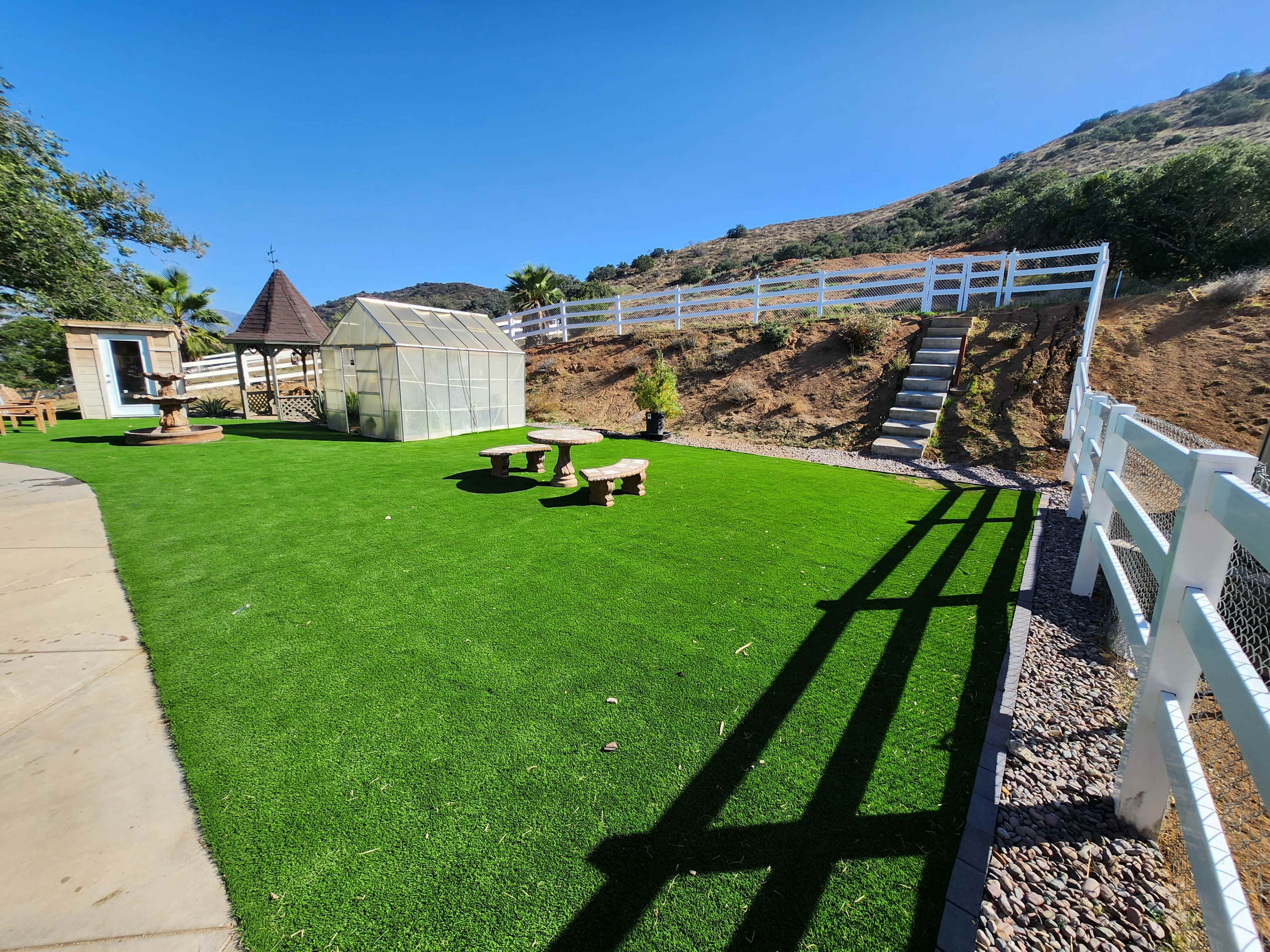 A landscaped yard with artificial grass, a greenhouse, a gazebo, and a staircase leading up a hillside.