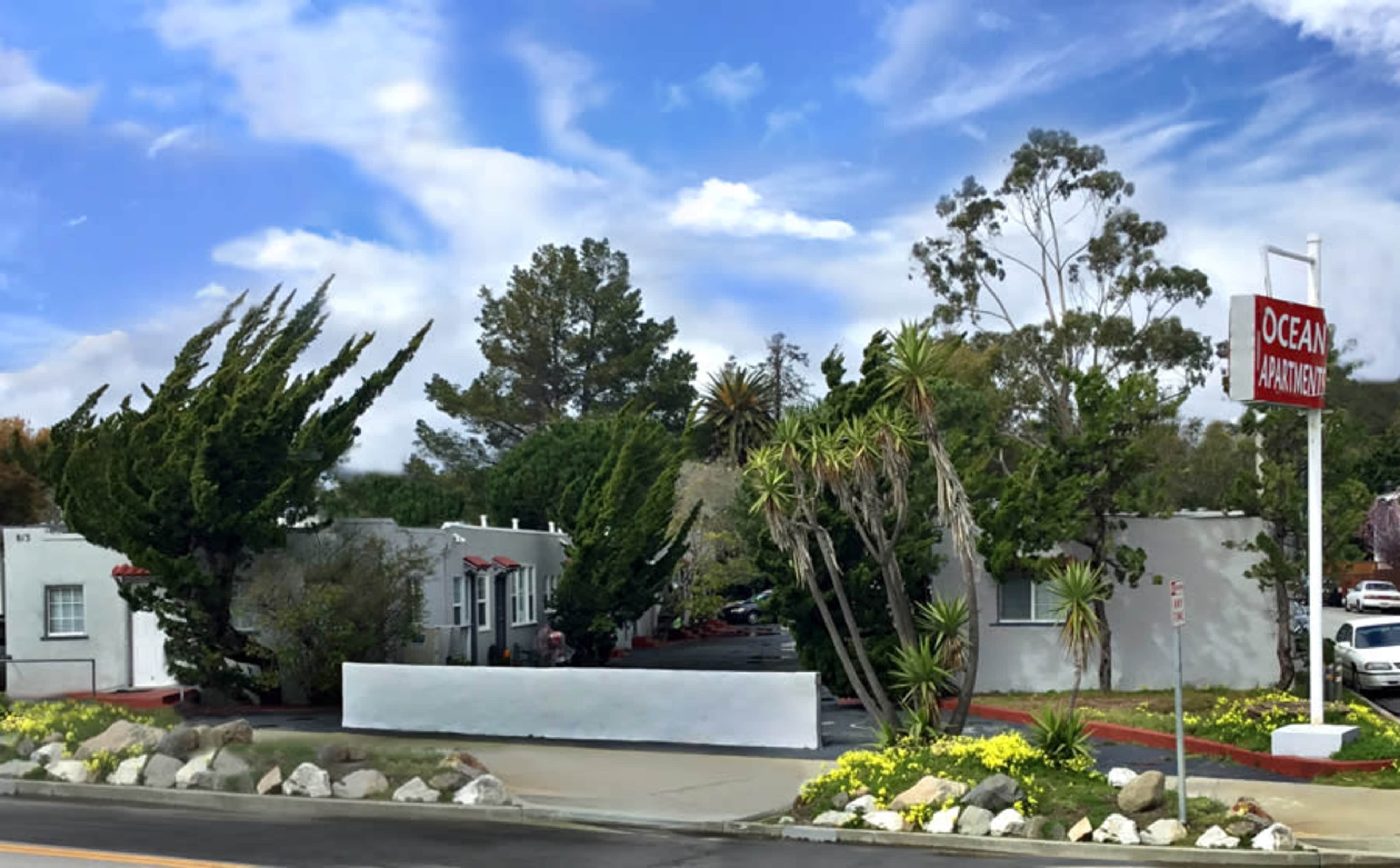 The image shows a row of apartment buildings with landscaped greenery and palm trees under a blue sky.