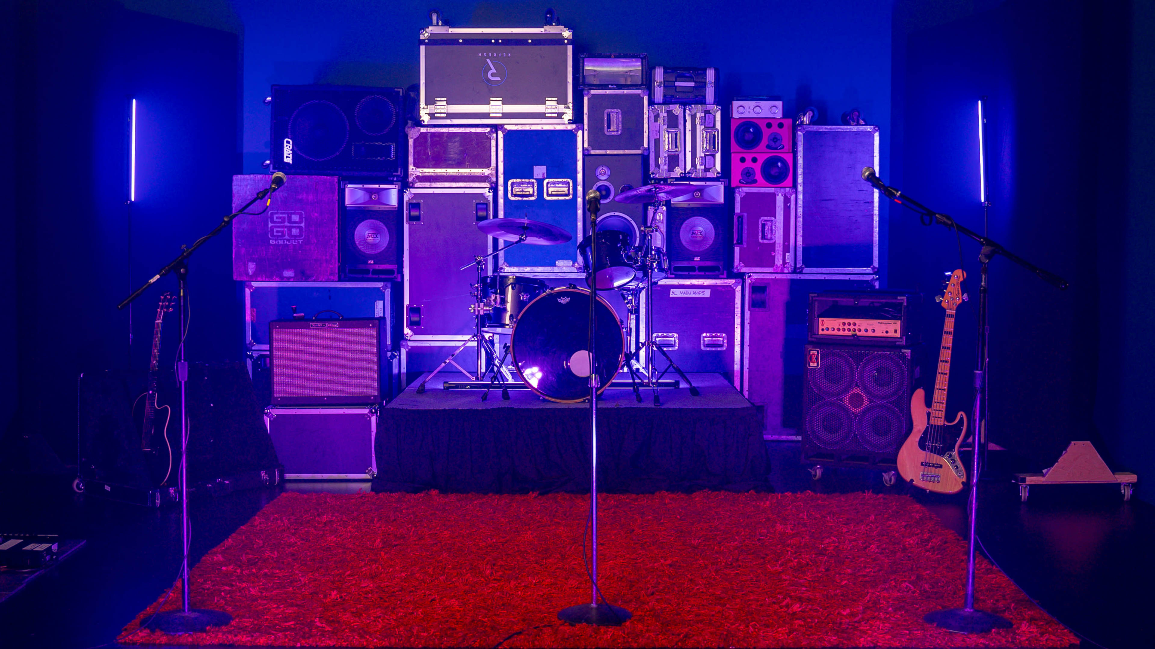 A stage setup features a drum kit surrounded by stacks of amplifiers and speakers, with three microphone stands in front and a red rug on the floor.