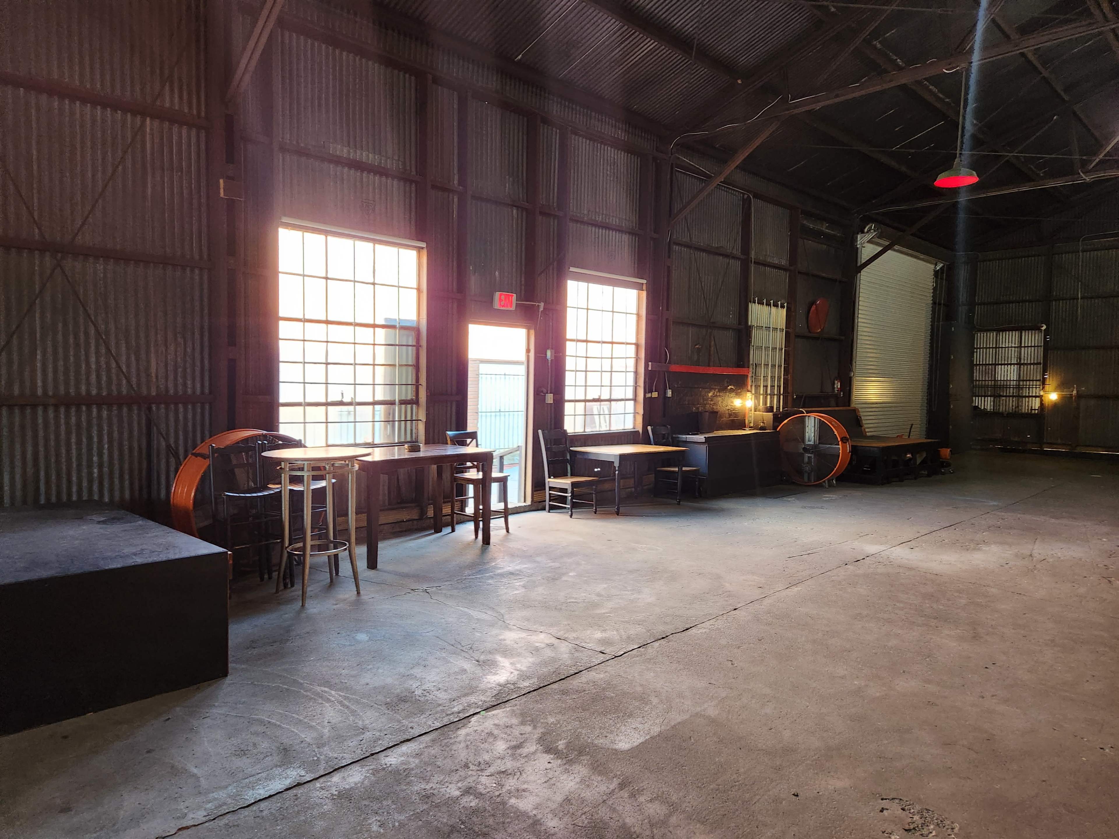 An industrial interior with metal walls, large windows, and sparse furniture, including tables and stools.