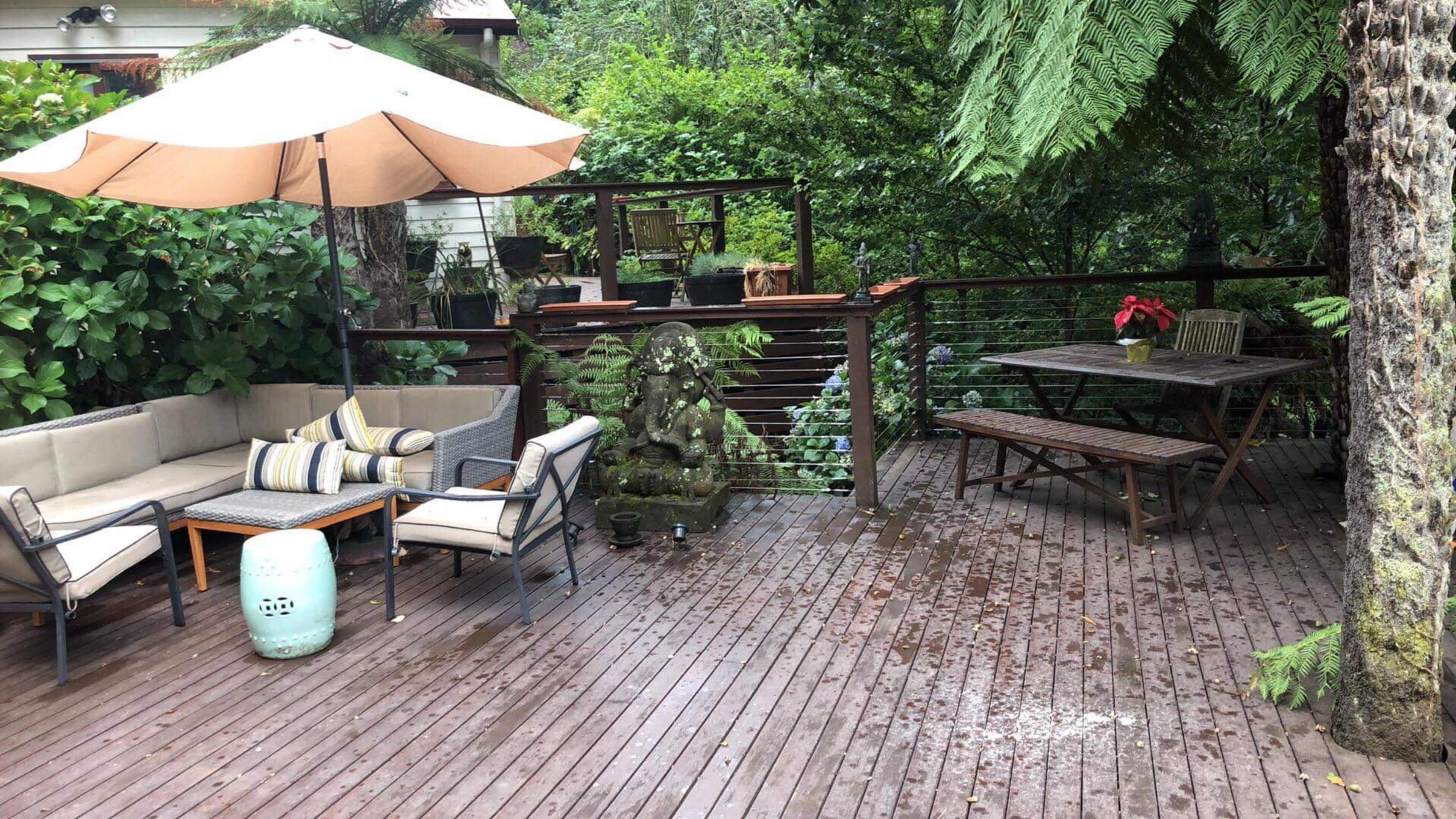 A wooden deck area with a seating arrangement under an umbrella, a small stone statue, and a dining table surrounded by lush greenery.