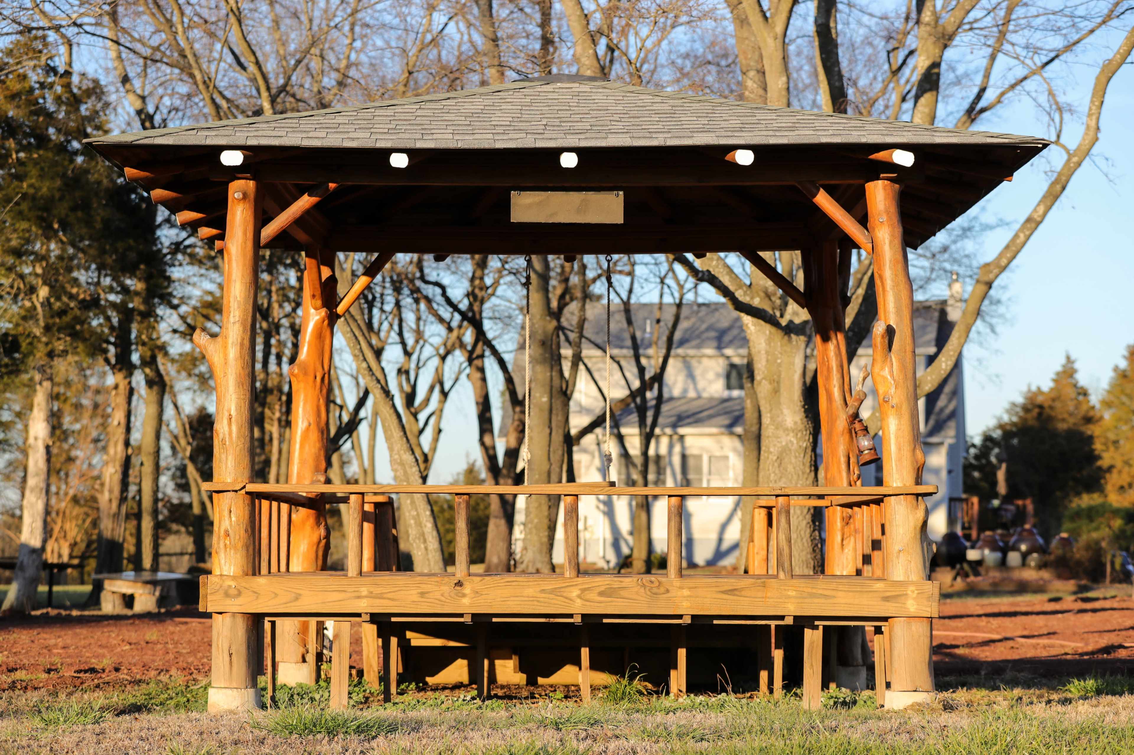A wooden gazebo stands in a park, surrounded by trees and a house in the background.