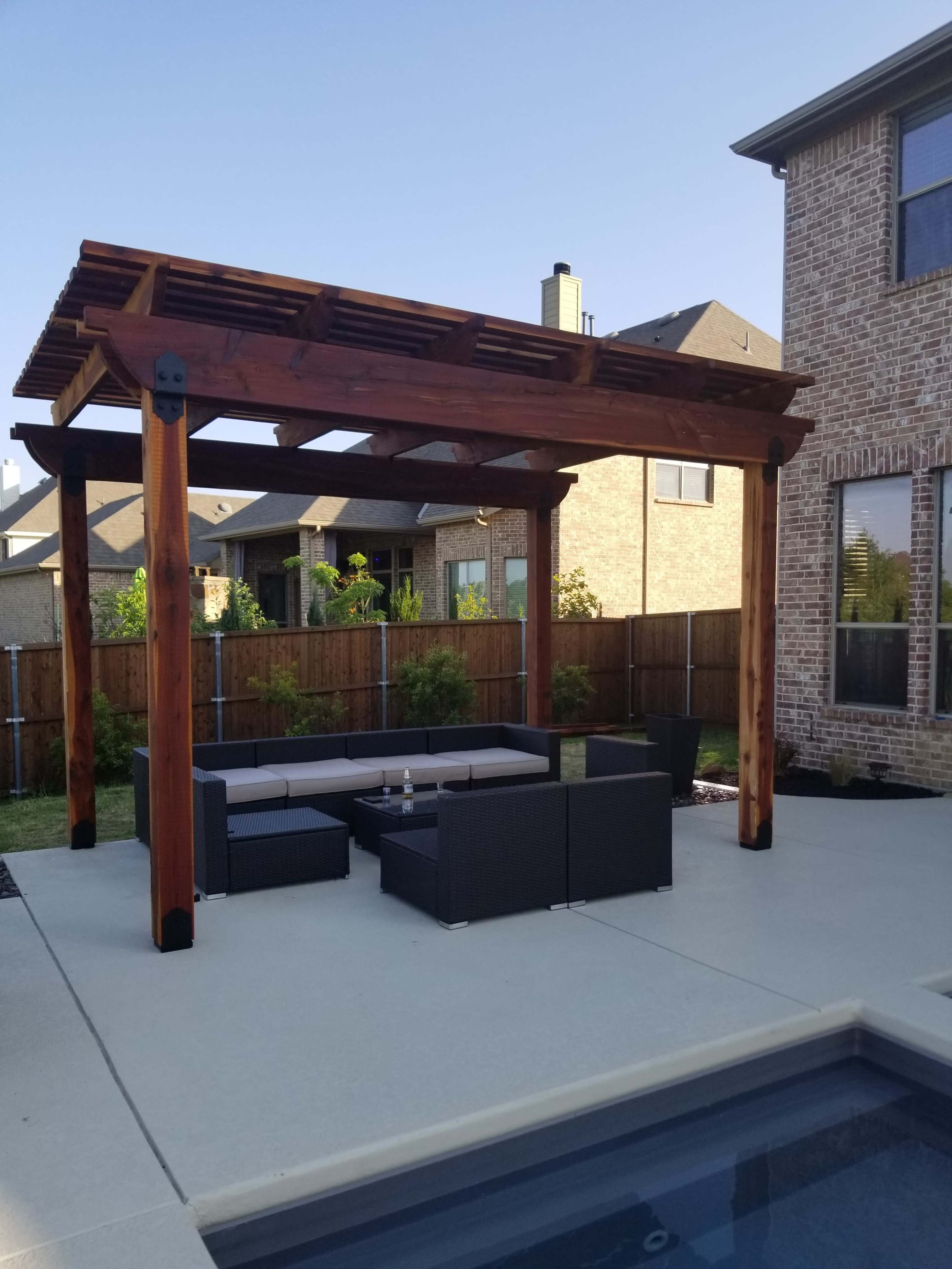 A wooden pergola stands over a seating area with black outdoor furniture next to a pool in a residential backyard.