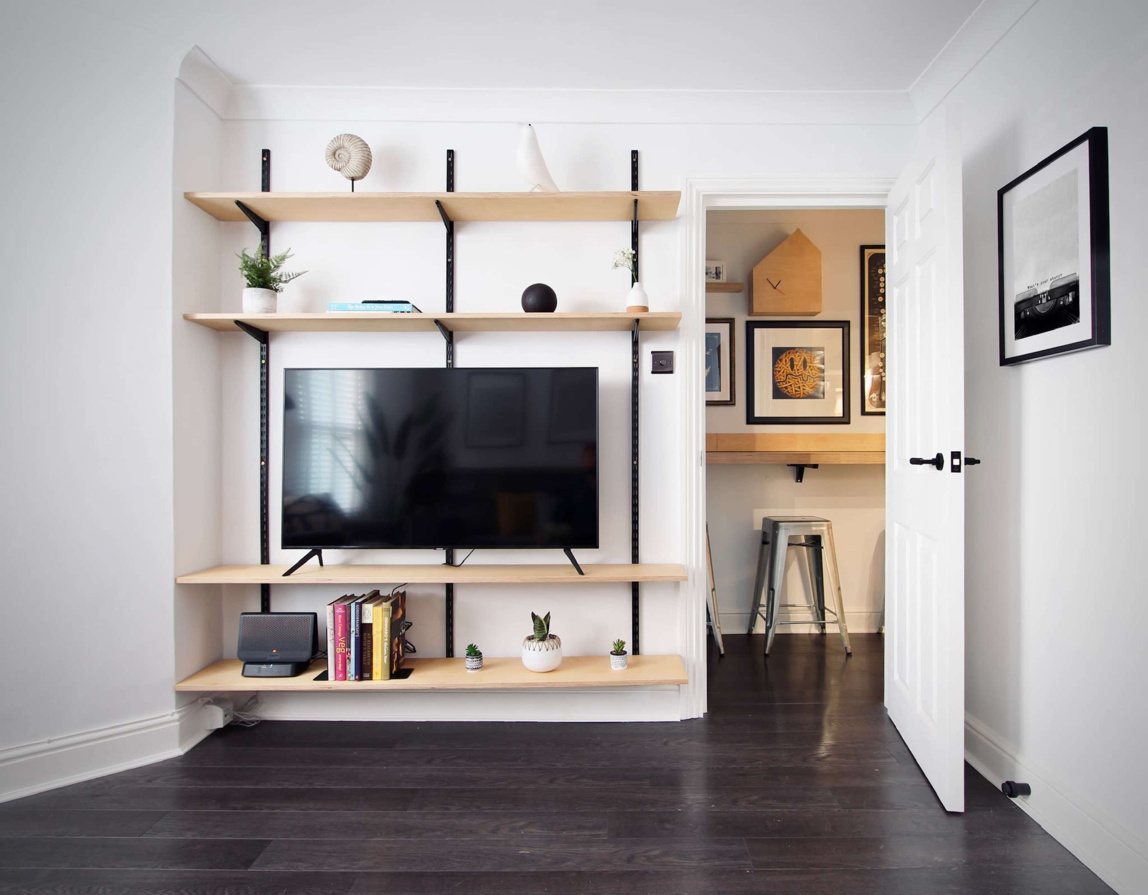 The image shows a modern living room with a wall-mounted television on a wooden shelving unit, surrounded by decorative plants and various books.