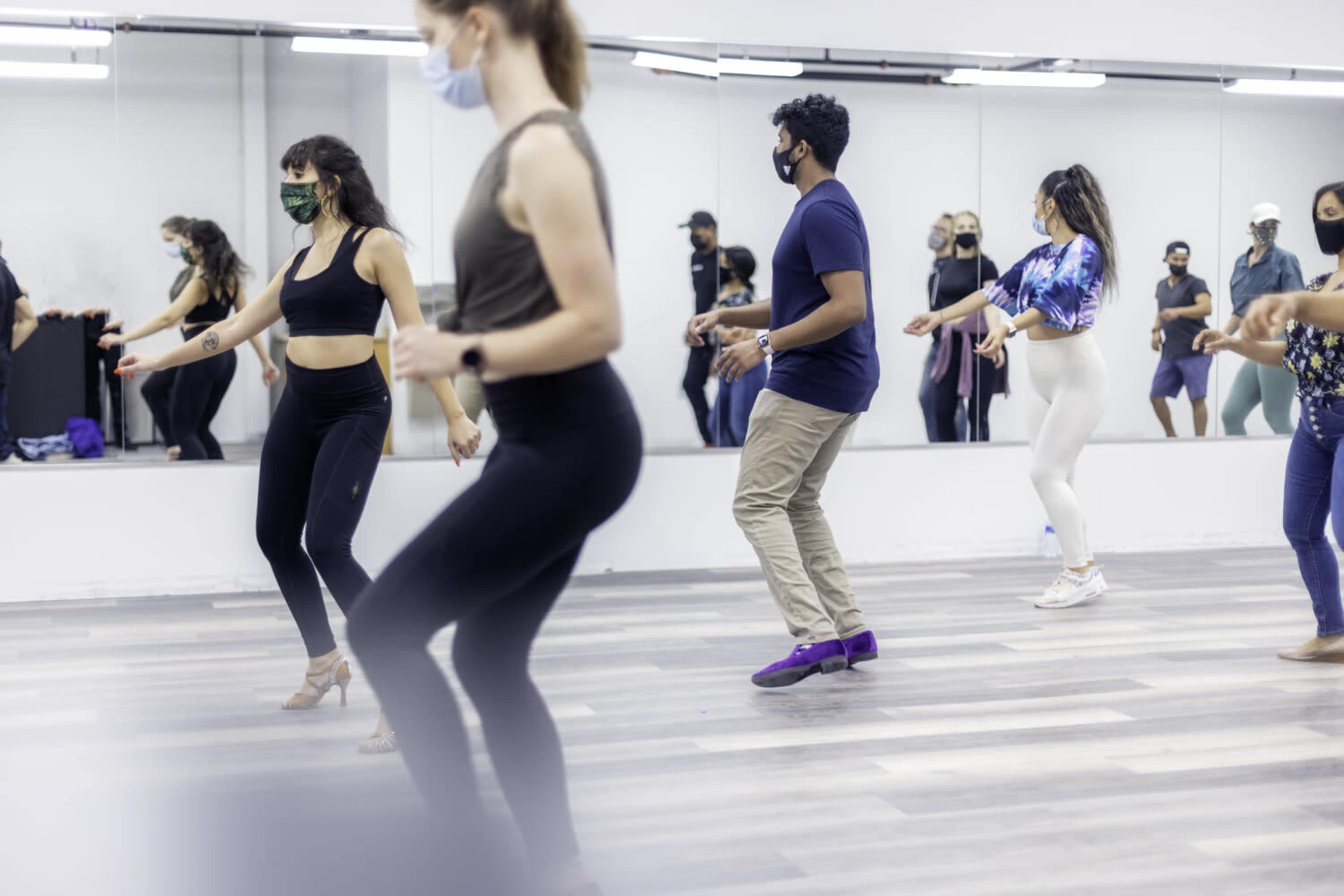 A group of people wearing masks are dancing in a studio with large mirrors.