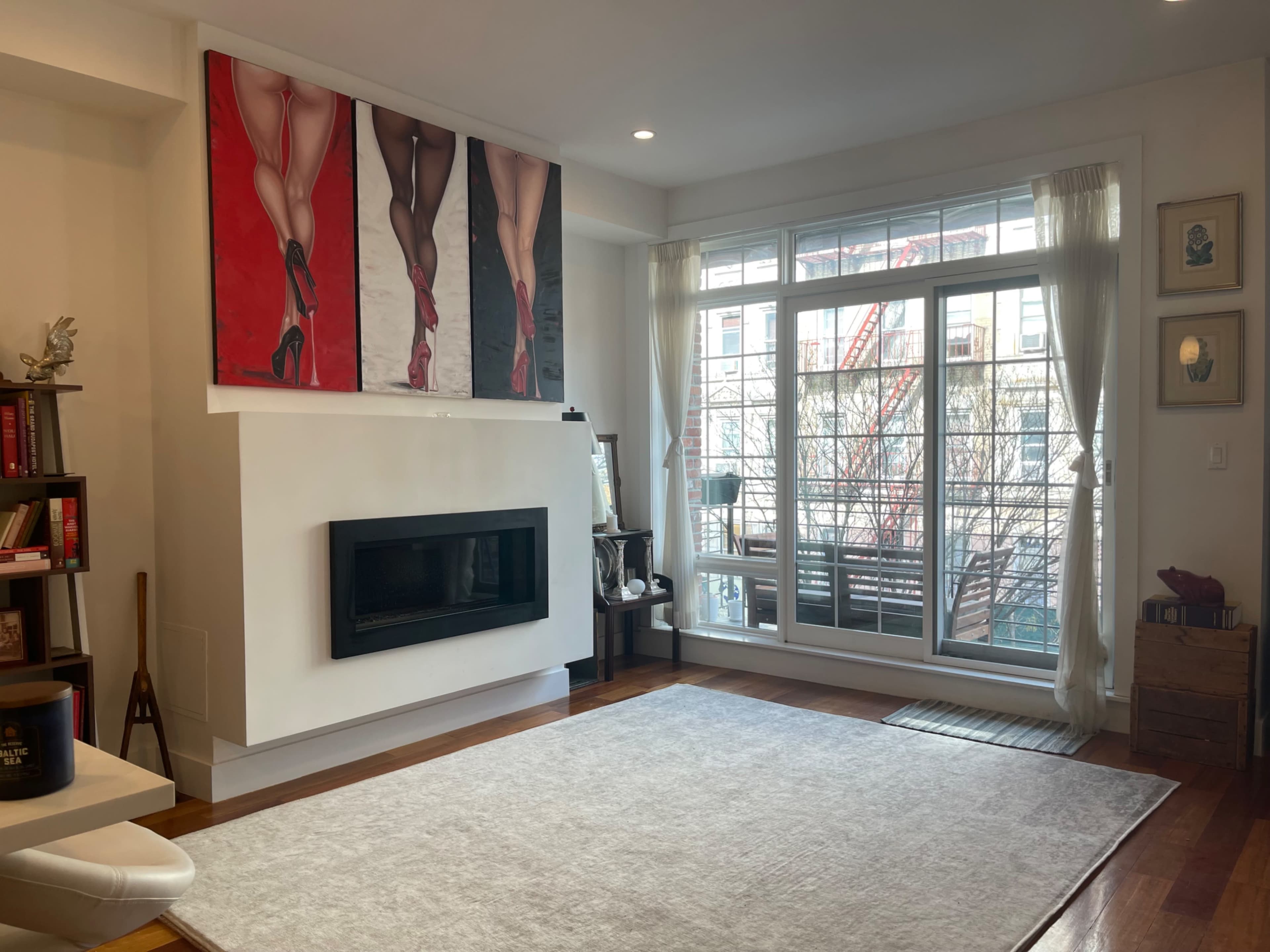 A modern living room featuring a fireplace, a large window with sheer curtains, and a wall adorned with a triptych of artwork depicting legs in high heels.