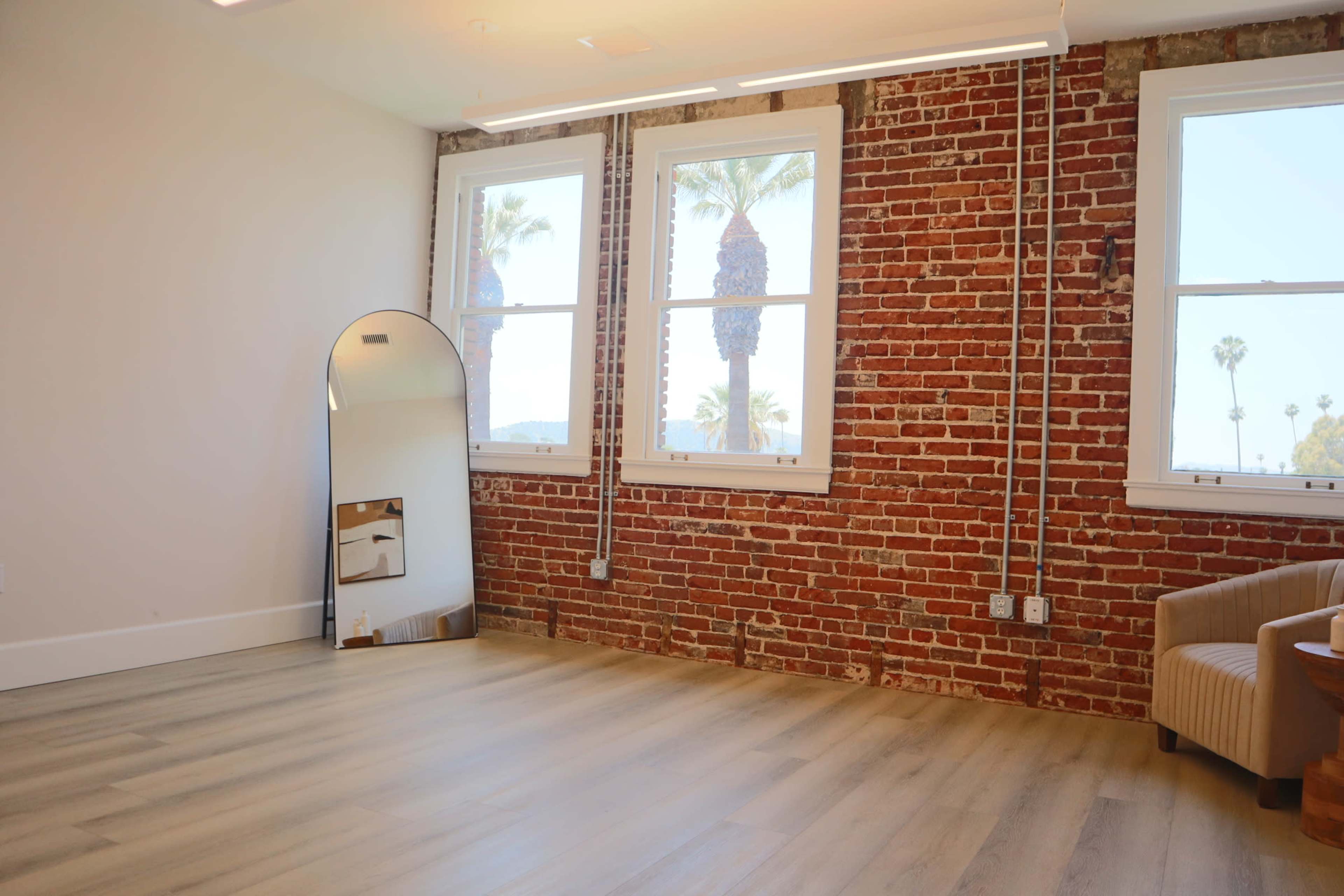 The room features exposed brick walls, large windows with palm trees visible outside, and a full-length mirror against the wall.