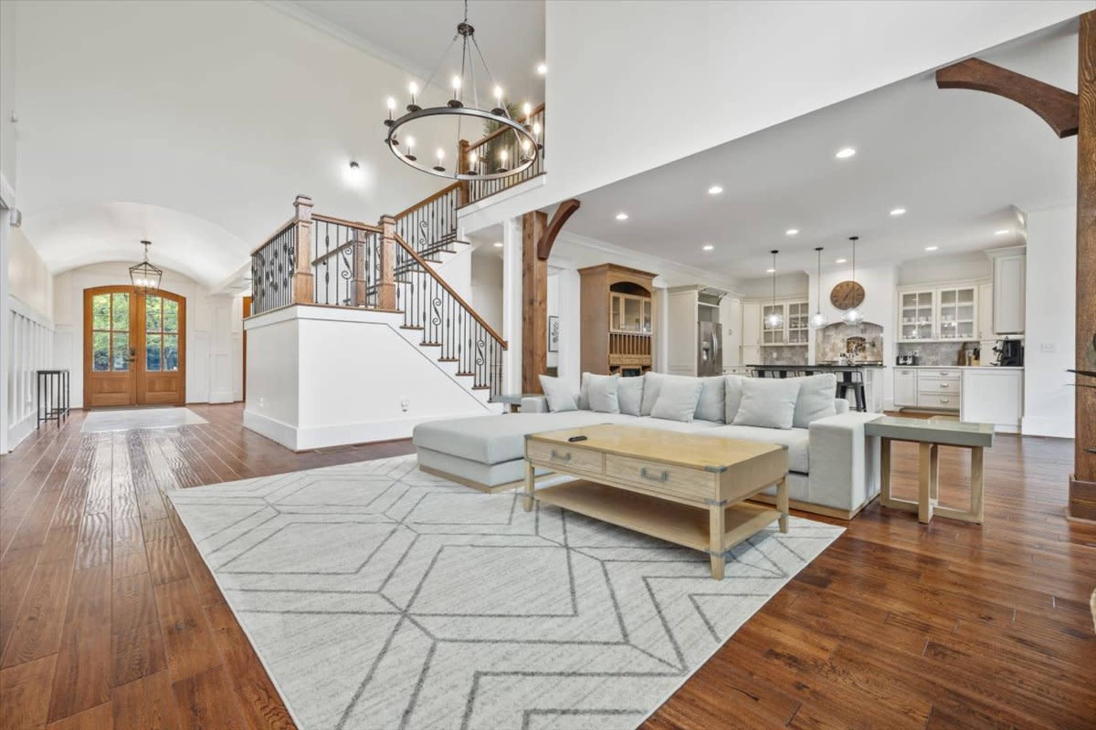 A spacious living area with a light gray sectional sofa, a wooden coffee table, and a large area rug, adjacent to a stairway and opening towards a kitchen area.