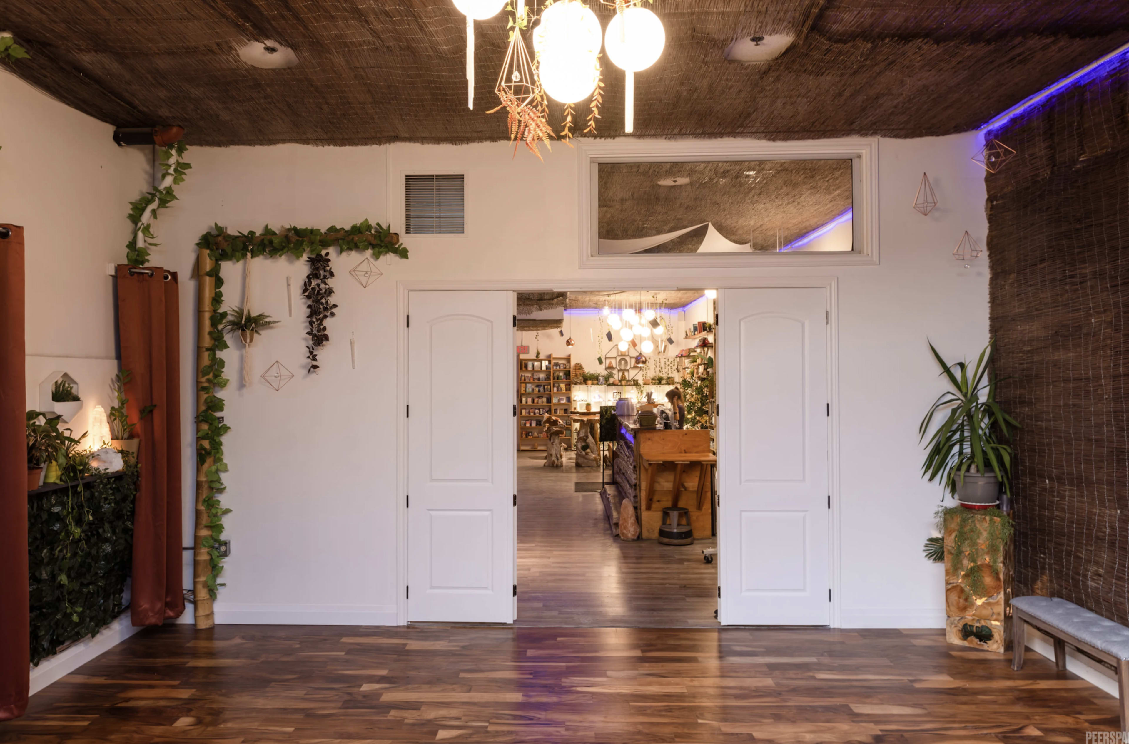 The image shows an interior space with a doorway leading to another room, featuring wooden flooring, wall plants, and decorative lighting.