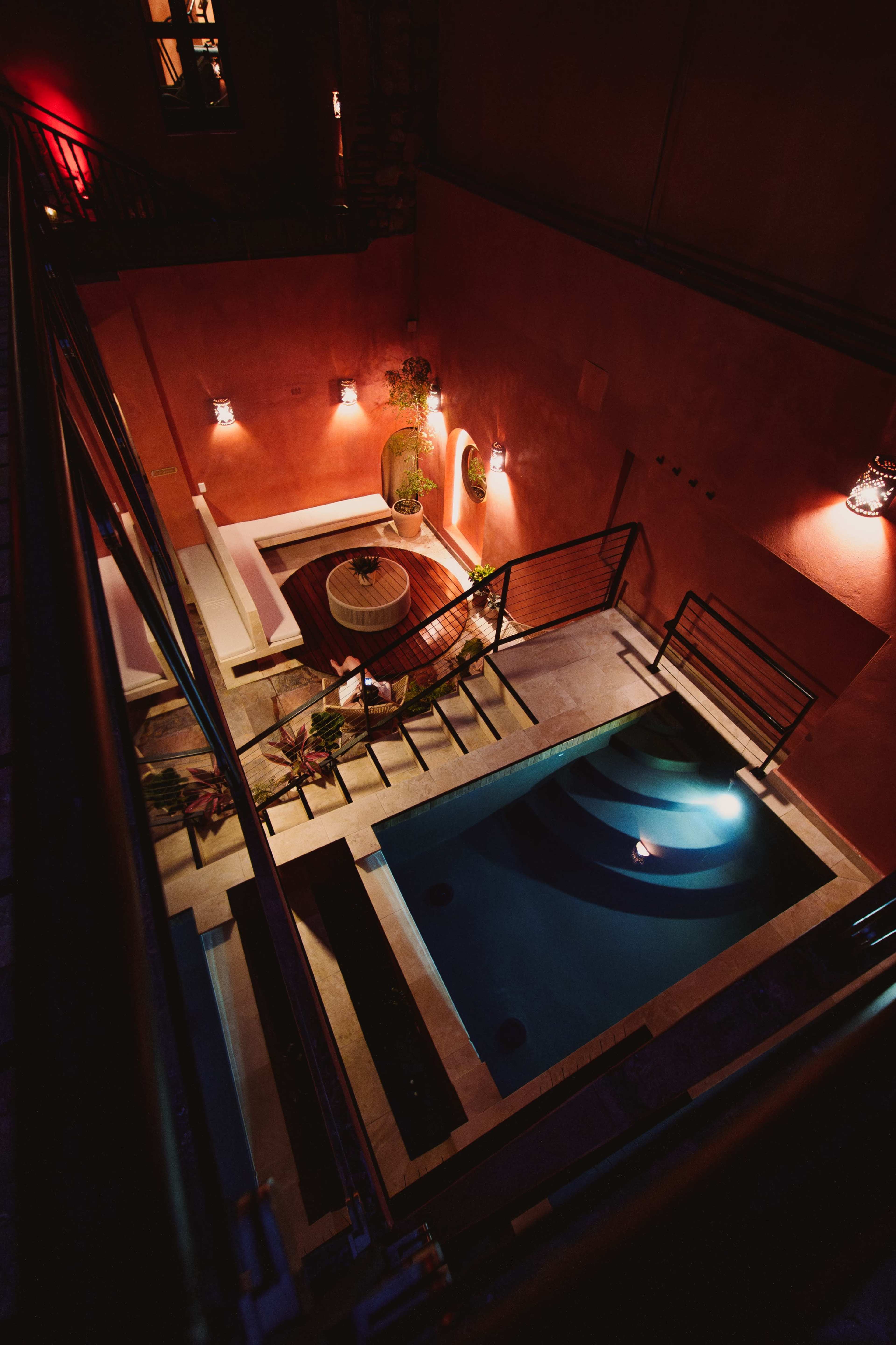 The image shows an aerial view of a modern courtyard featuring a rectangular swimming pool surrounded by plants and illuminated by wall sconces.