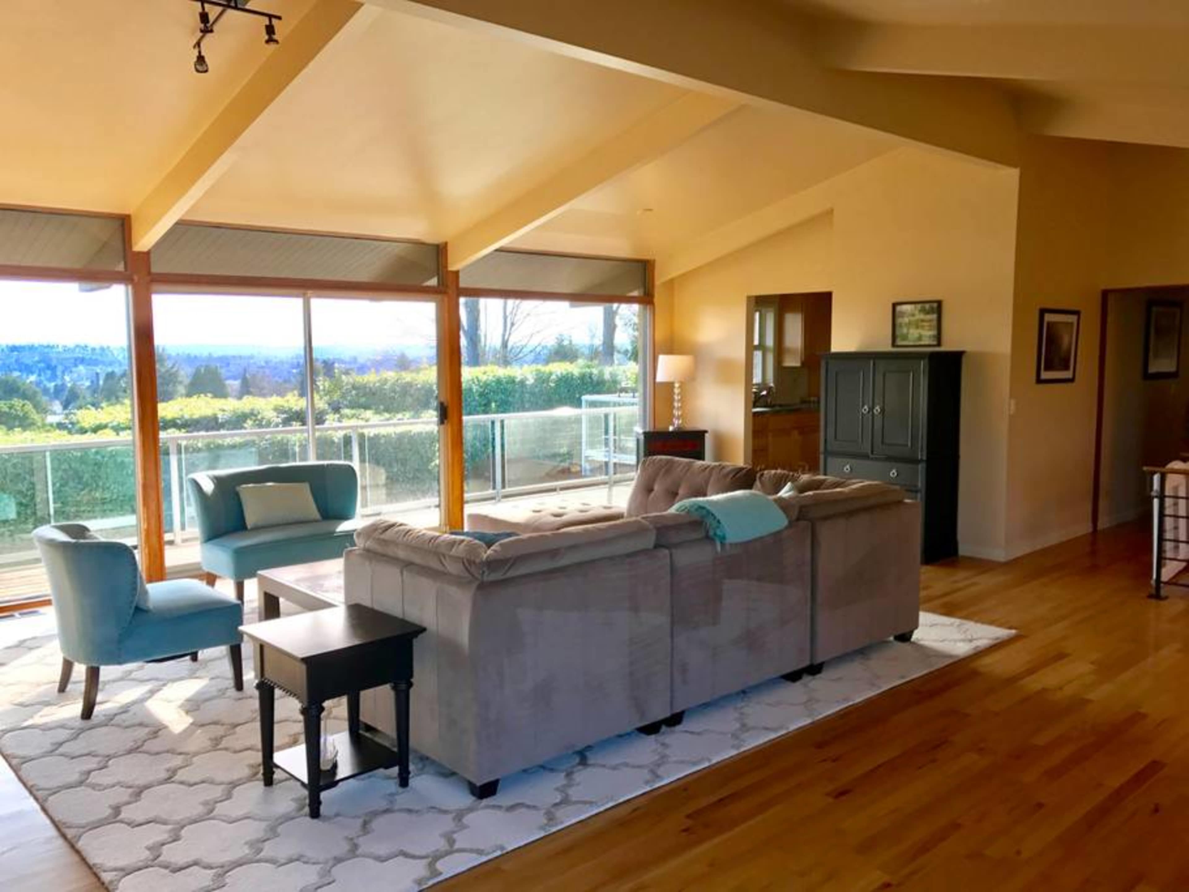 The living room features a sectional sofa, two blue armchairs, and large windows that overlook a scenic view.
