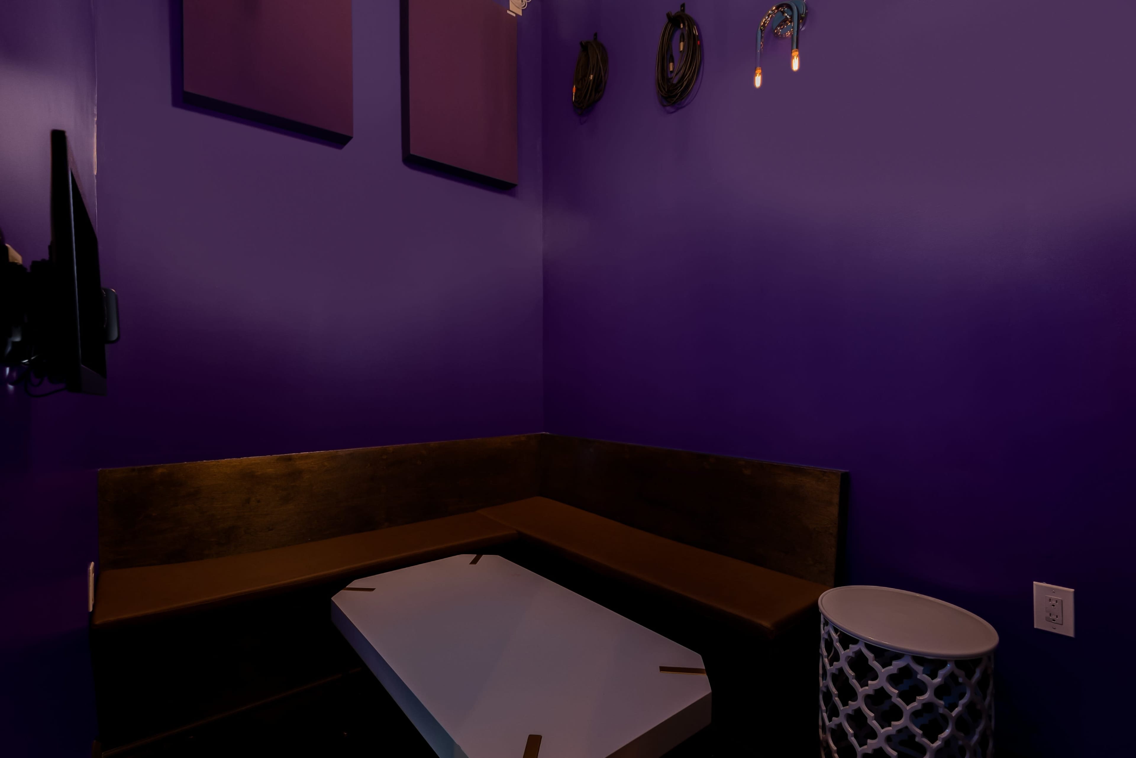 The image shows a small purple-walled room featuring a seating area with a wooden bench, a white table, and decorative wall elements.
