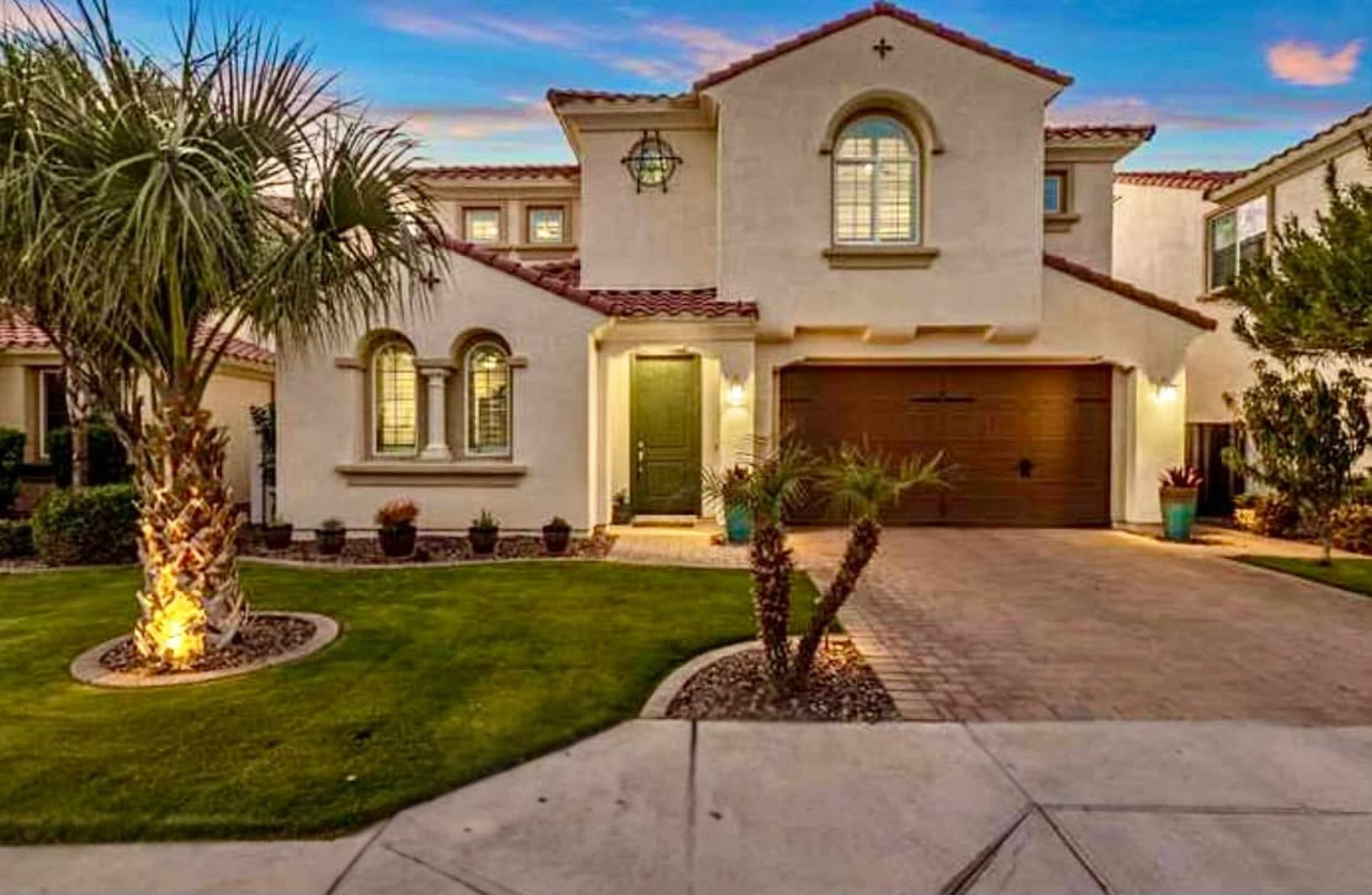 A stucco home with a tiled roof stands beside a manicured lawn and palm trees, featuring a two-car garage and a stone pathway.