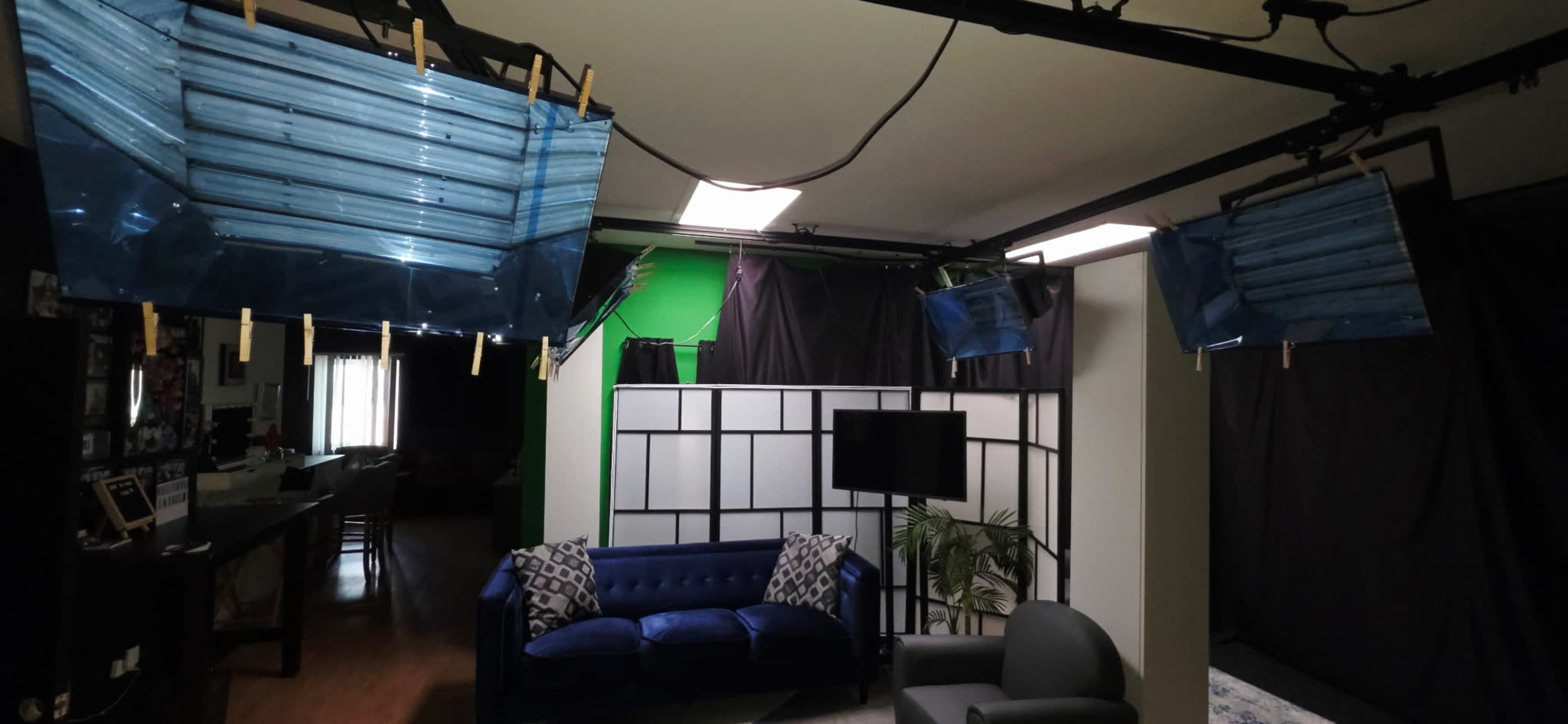 The image shows a room set up for filming, featuring blue lighting panels suspended from the ceiling, a blue couch, a television on a stand, and a green screen backdrop.