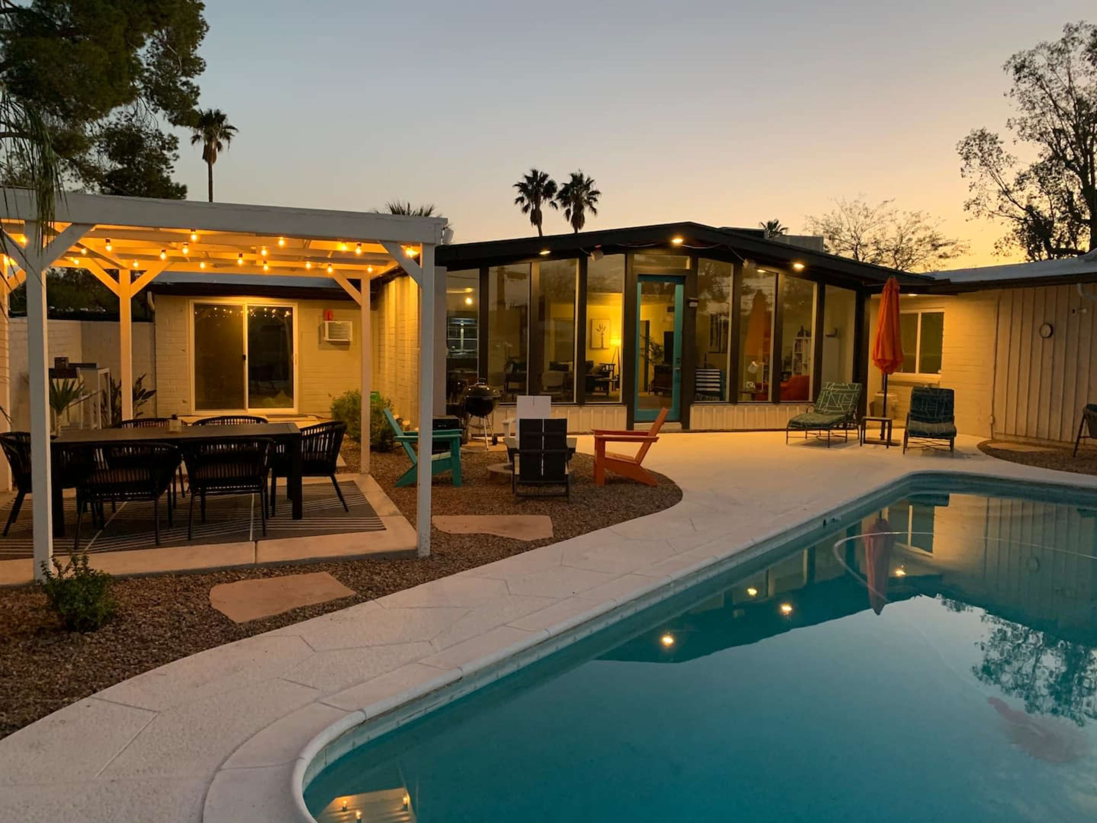 A modern backyard with a swimming pool, a covered dining area with string lights, and a single-story house surrounded by palm trees at dusk.