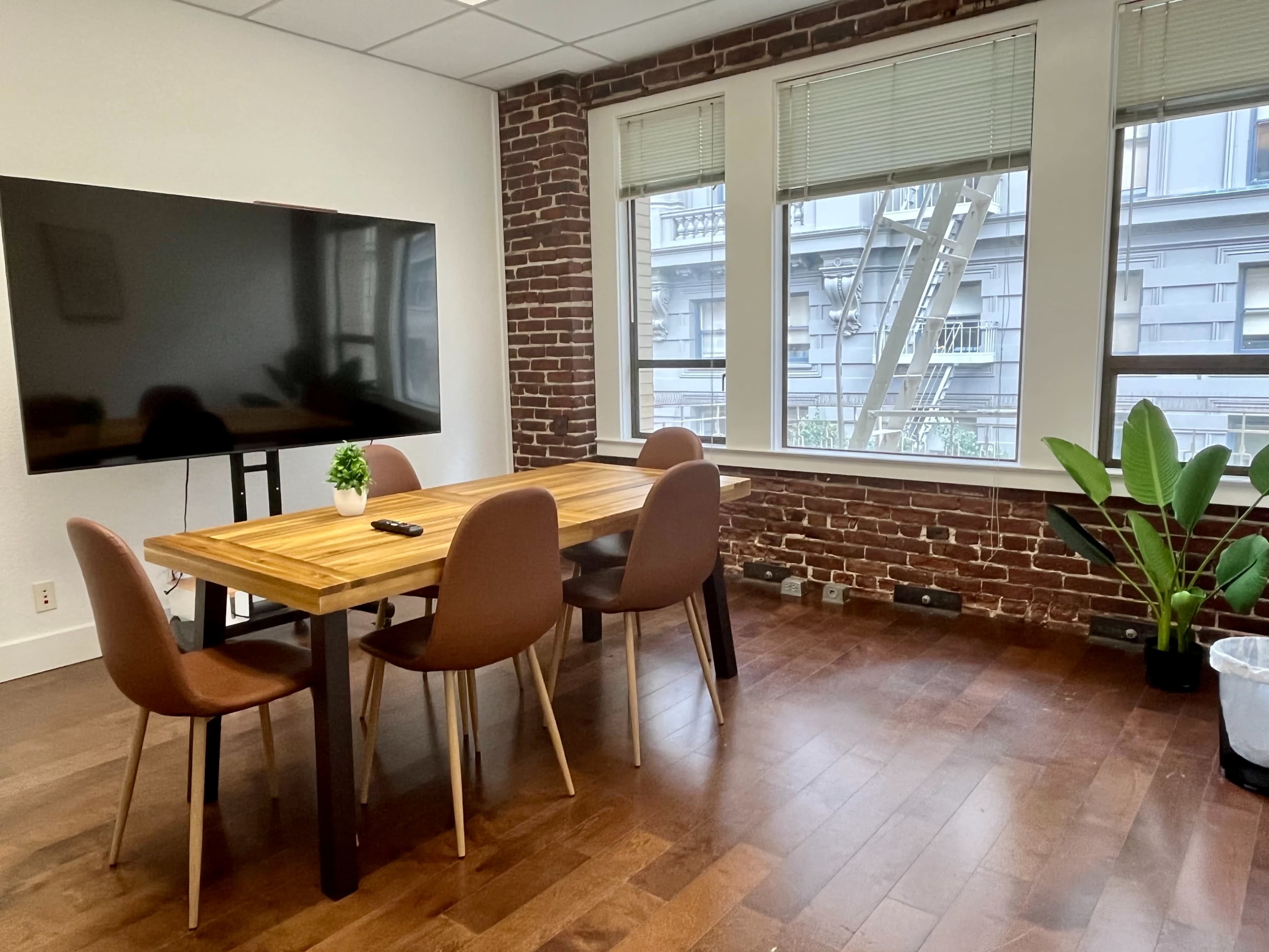 A meeting room features a wooden table surrounded by four chairs, with a large screen on a stand and large windows that offer a view of brick buildings outside.