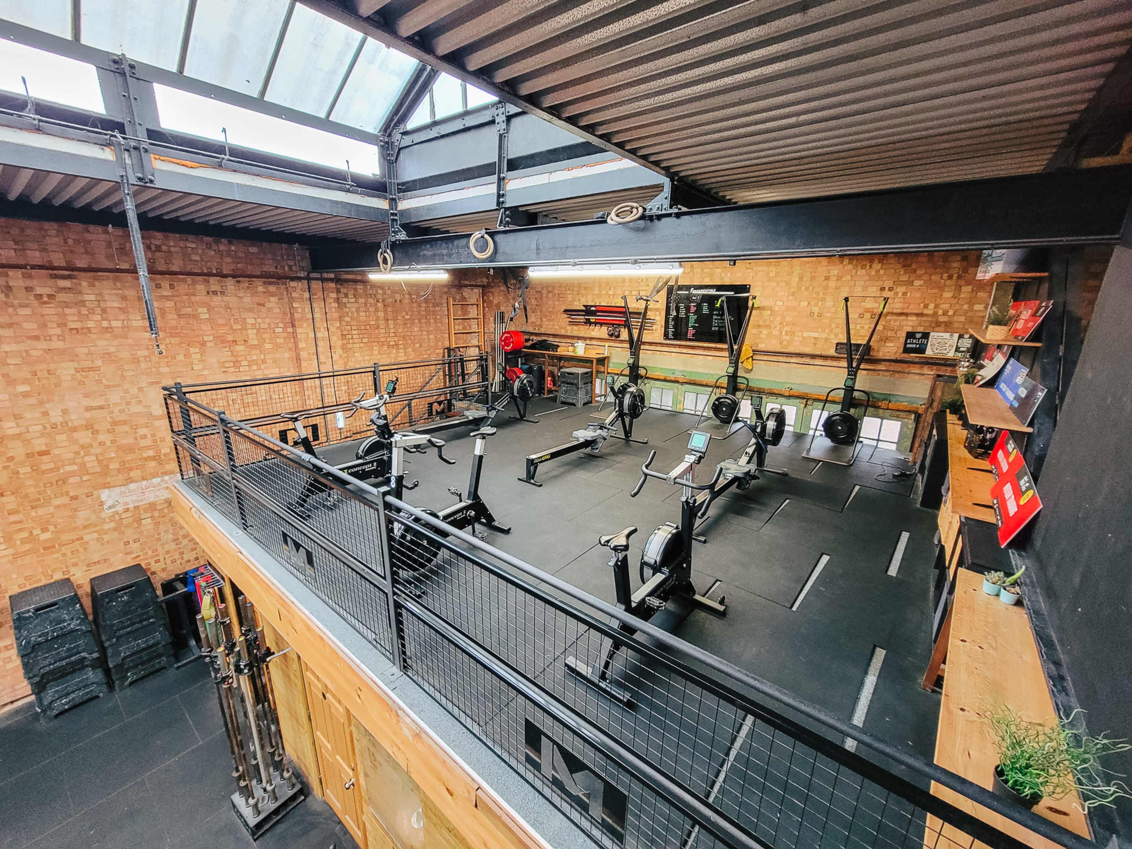 The image shows an indoor gym with a multi-level layout, featuring workout equipment including rowing machines and weightlifting benches against a brick wall.