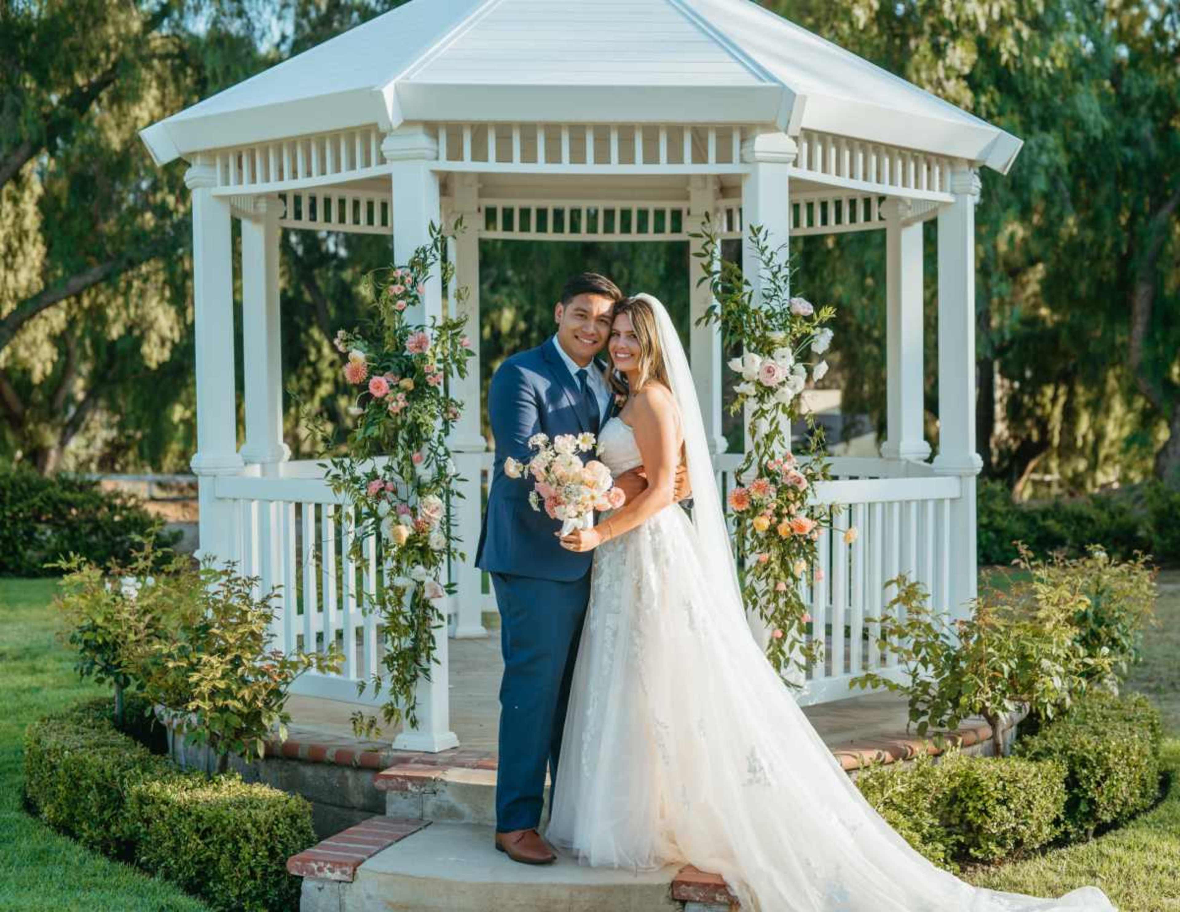 A couple stands together in front of a decorated gazebo, with flowers and greenery around them, as they hold a bouquet.