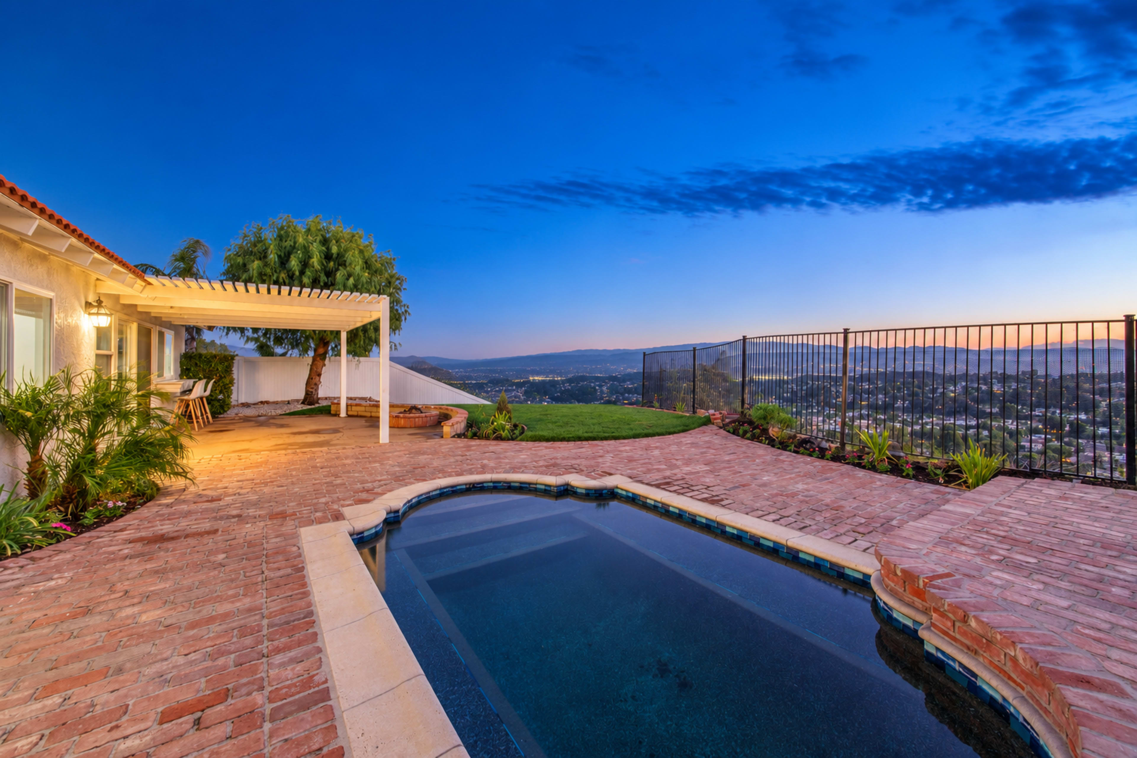 A backyard with a small pool, surrounded by brick paving and overlooking a scenic view, features a pergola and lush greenery under a twilight sky.
