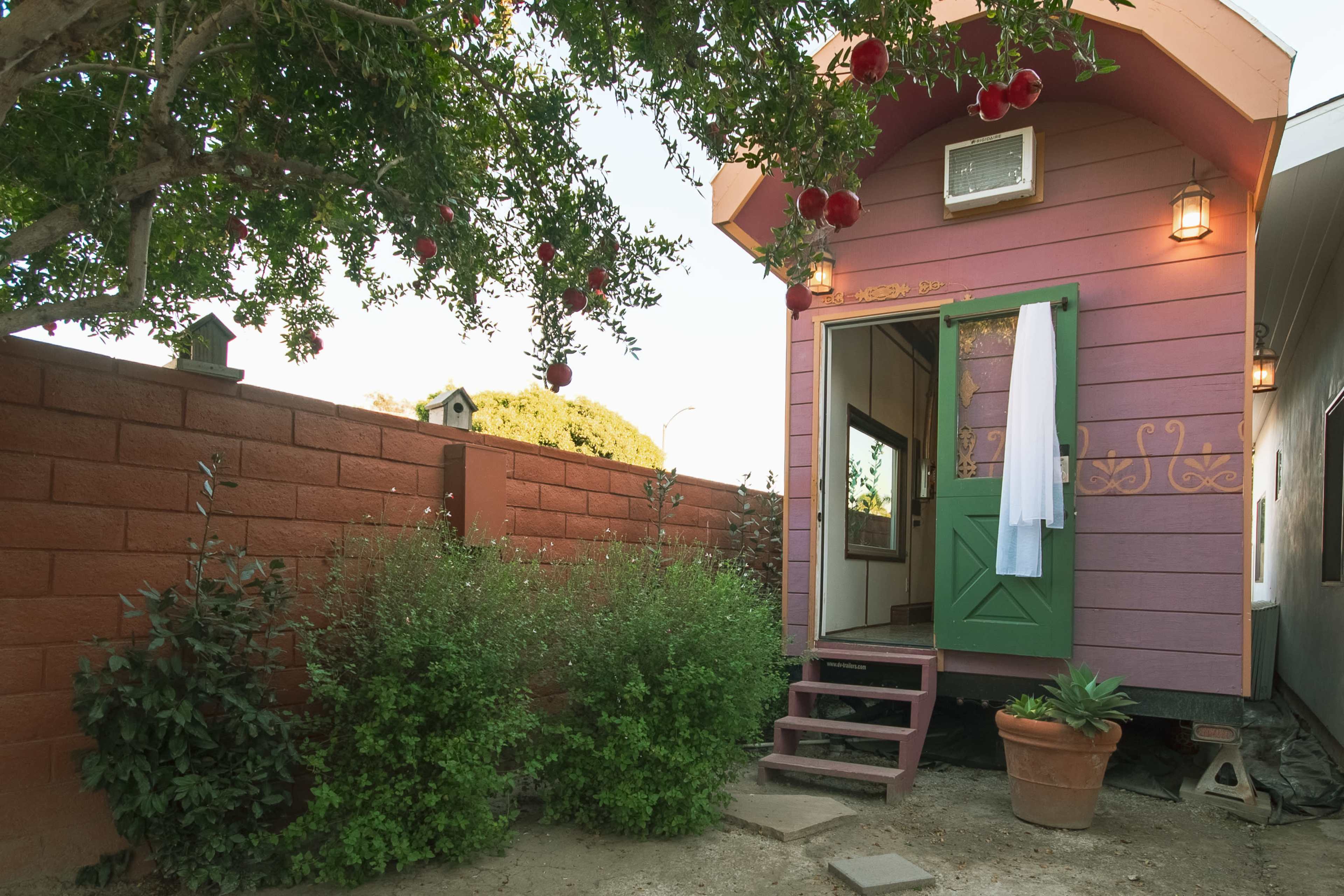 A small, colorful house with a partially visible door and window is situated next to a brick wall and surrounded by greenery and potted plants.